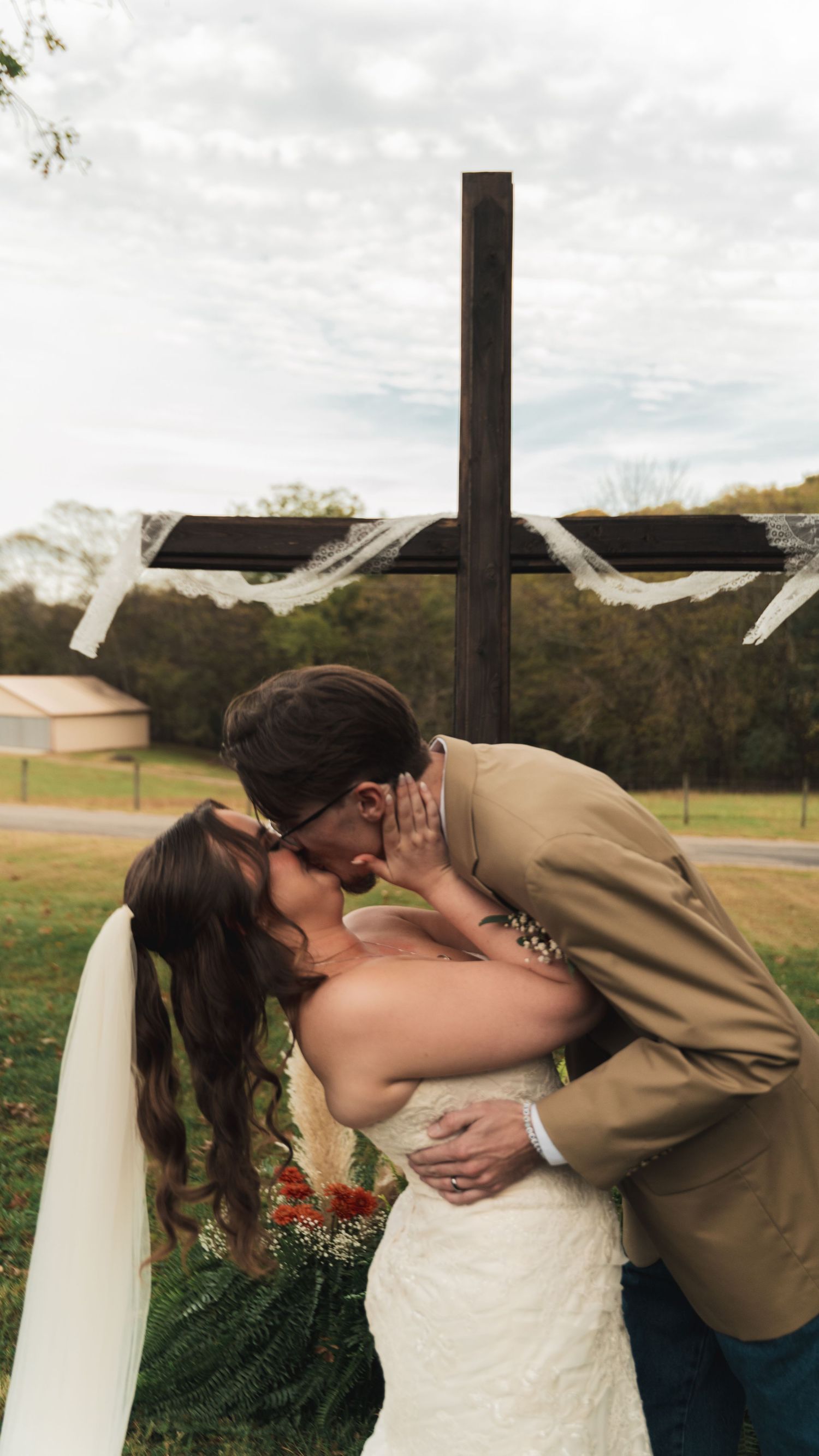Bride and groom kissing in front of a wooden cross at an outdoor wedding ceremony.