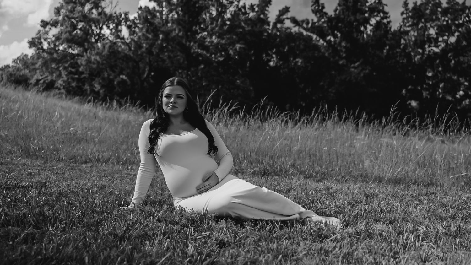 Pregnant woman in white outfit sitting gracefully on green grass field in black and white photography.