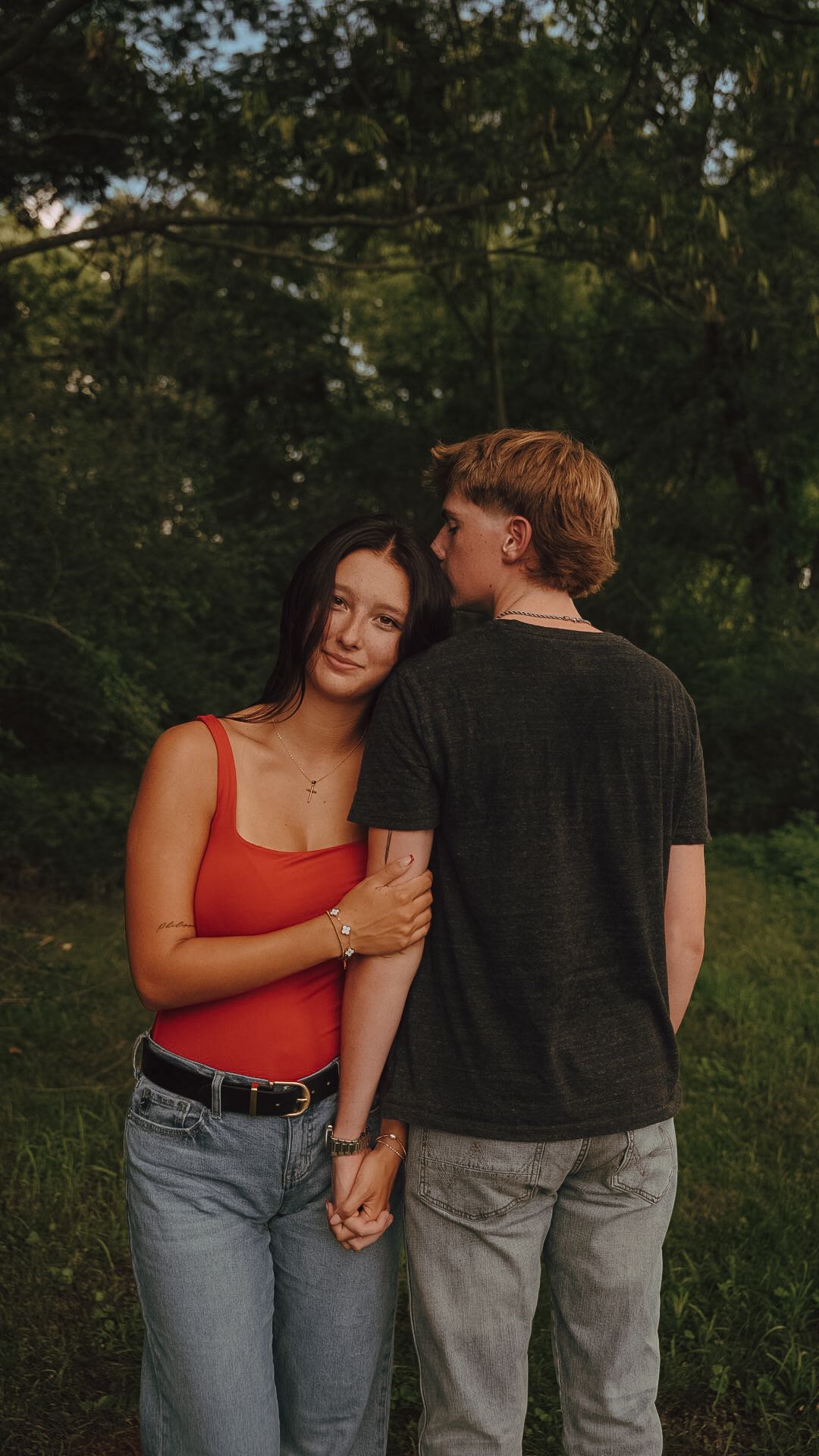 A young couple holds hands in a lush green park, the girl in a red tank top smiles as her partner embraces her.