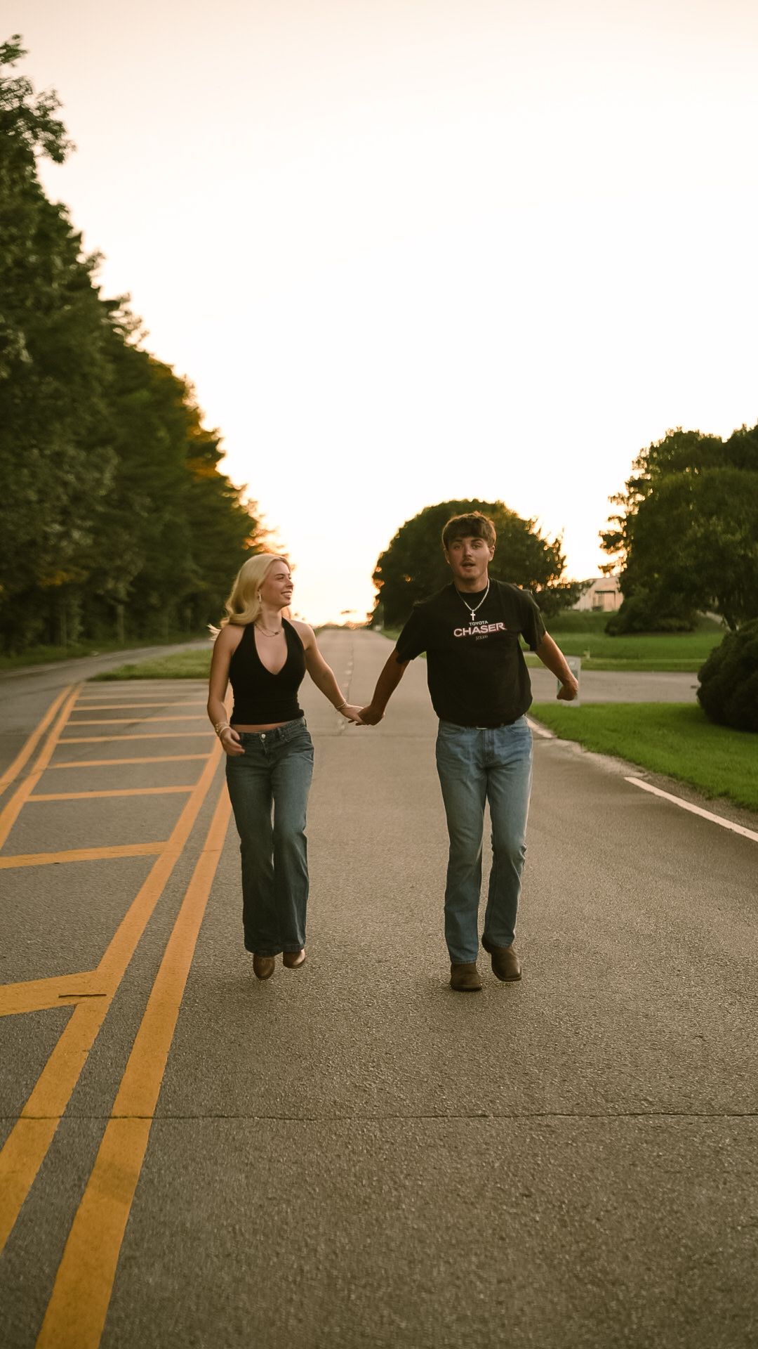 A couple holding hands and walking together on a tree-lined road at sunset, both wearing jeans and black tops.