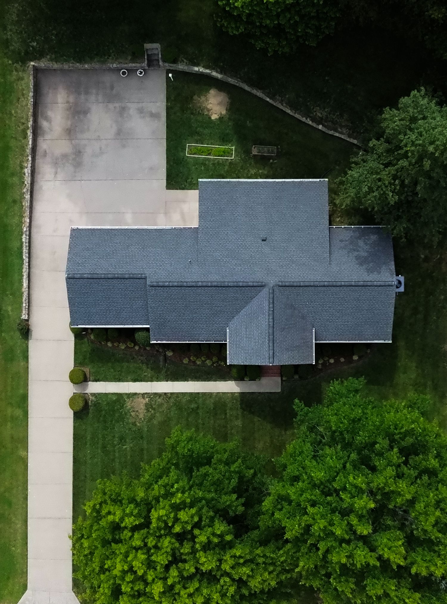 Aerial drone view of a suburban home with gray roof, concrete driveway, and lush green trees surrounding the property.