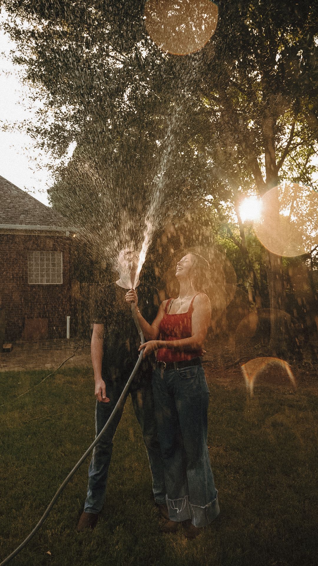 Couple laughing and playing with a garden hose in a sunny backyard, water spraying in golden sunset light.