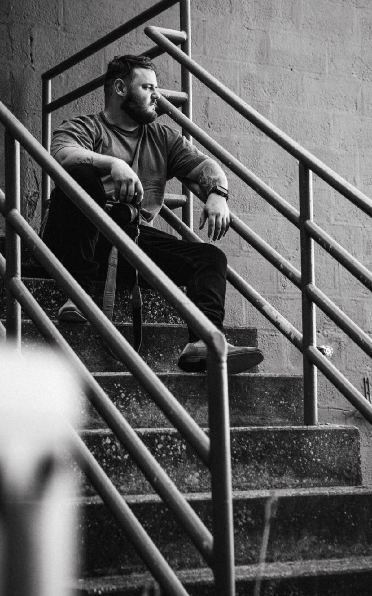 Black and white portrait of bearded man sitting on outdoor concrete staircase with railings.