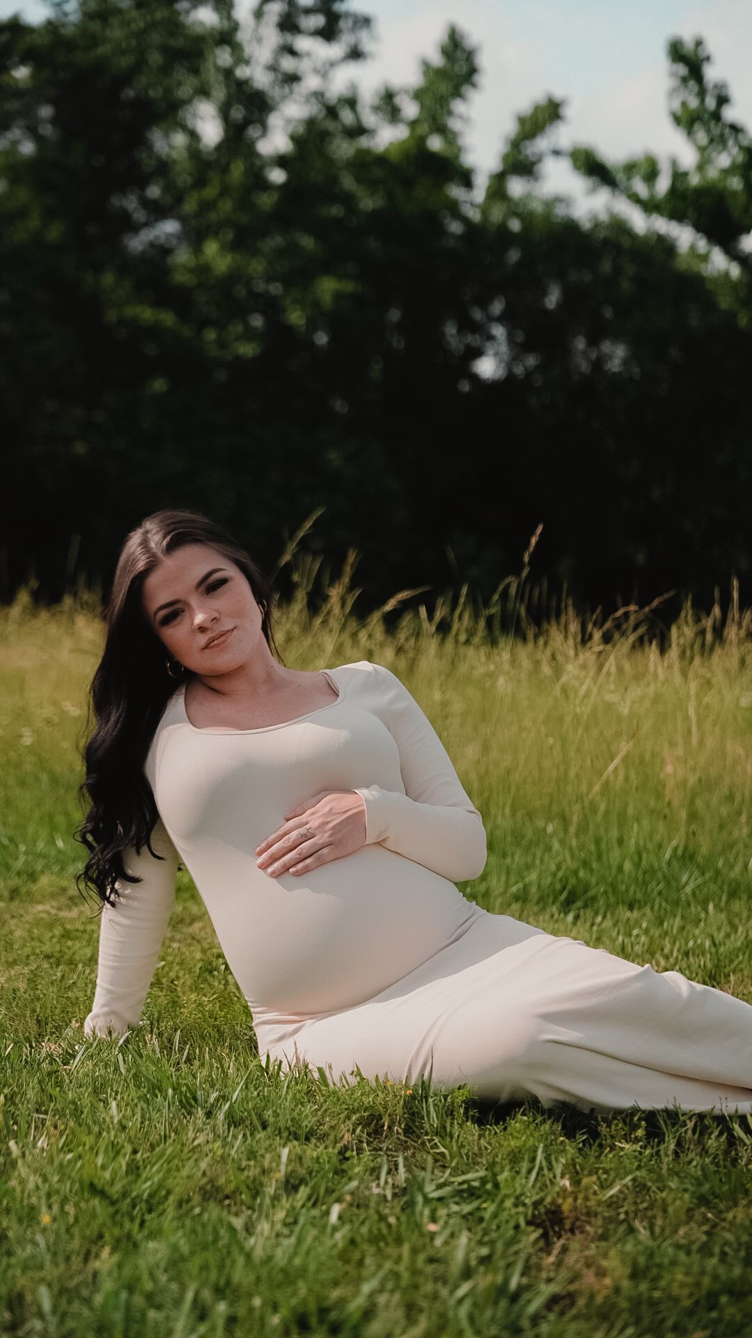Pregnant woman in white dress sitting on grass in a field, gently cradling her baby bump.