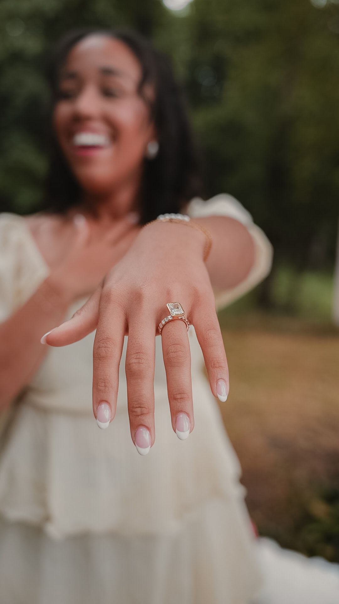 Smiling woman in white dress showing off her emerald cut diamond engagement ring with French tip nails outdoors.