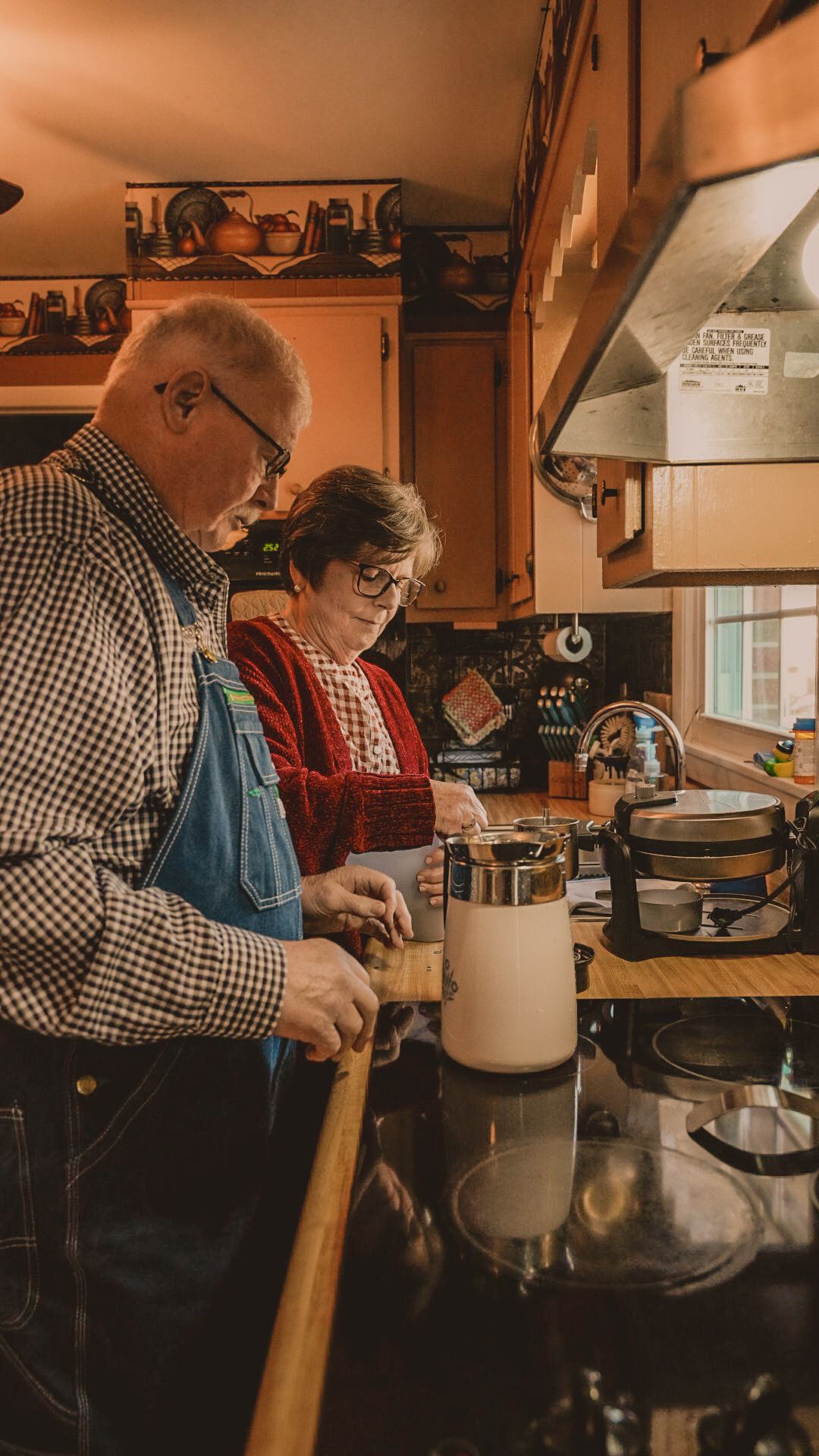 Elderly couple cooking together in a warm, cozy kitchen, man in overalls and woman in red apron preparing food at the counter.