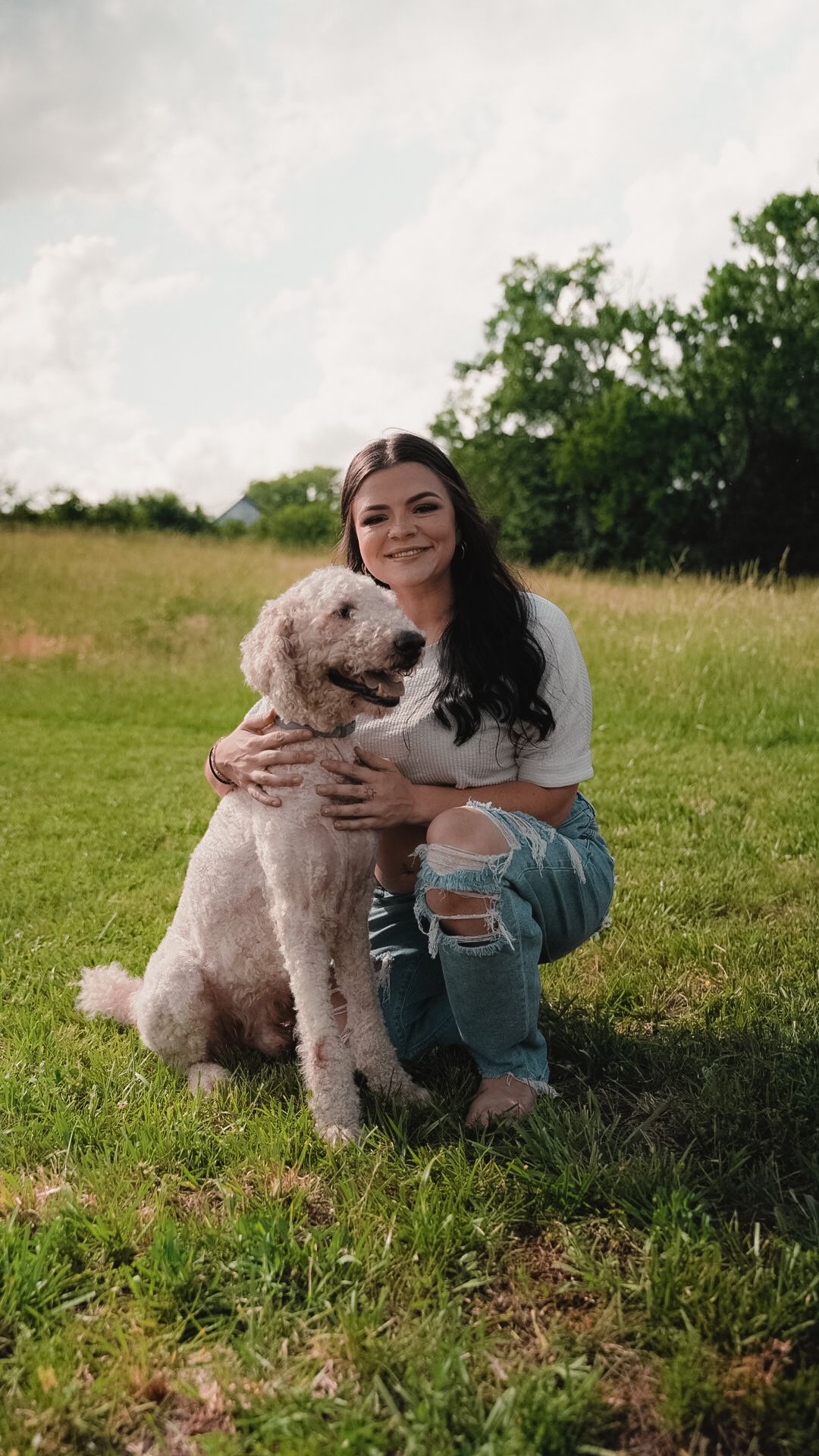 Young woman with dark hair smiling while hugging a fluffy goldendoodle dog on a green grass field.