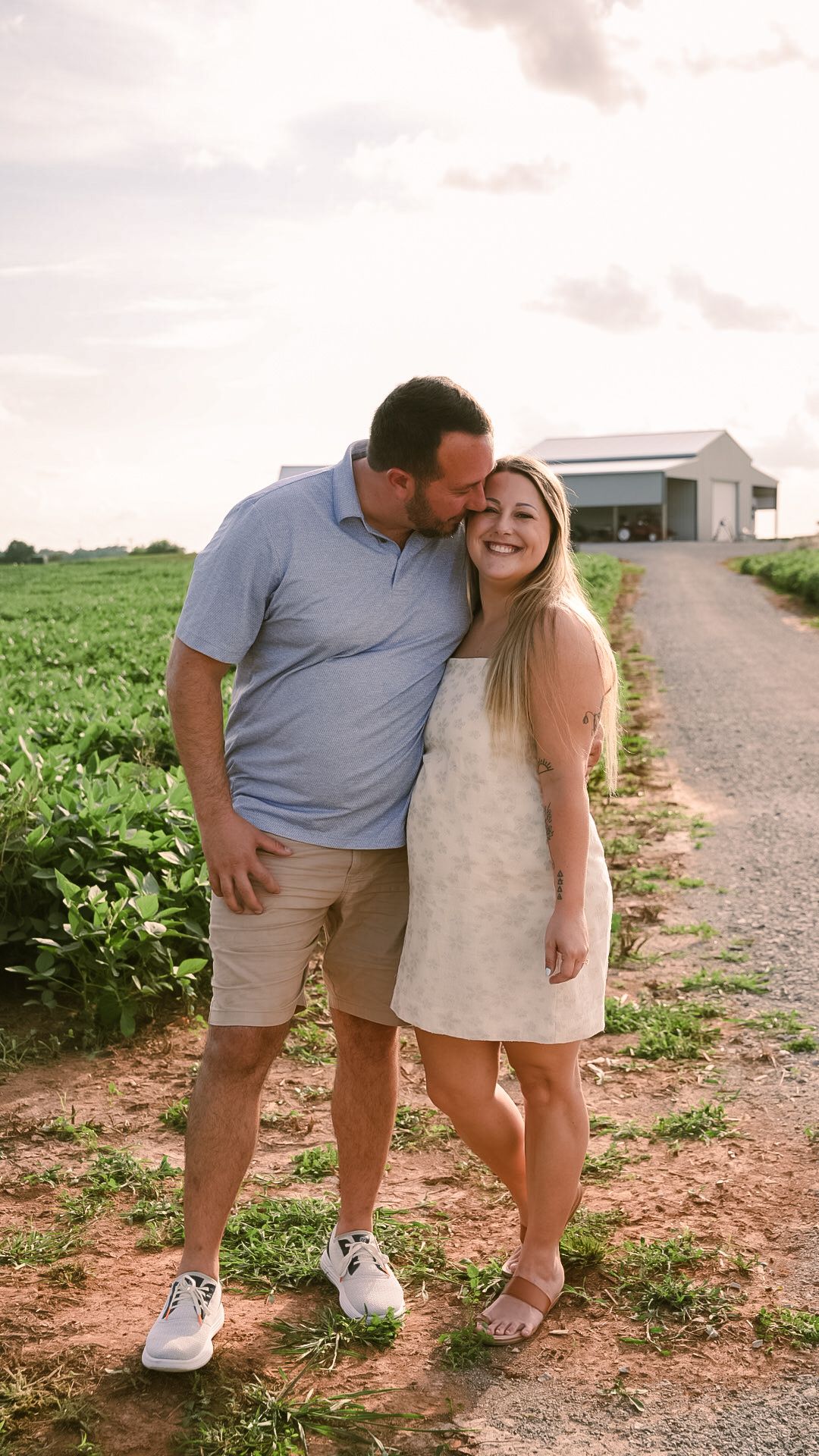 A couple poses on a farm path at sunset, man kissing woman's head, green fields and white barn behind them.