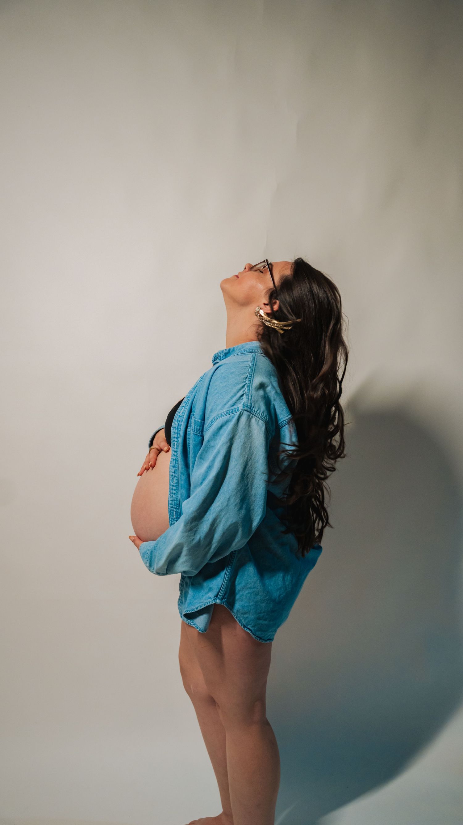 Pregnant woman in blue shirt cradling baby bump, head tilted back, long dark hair, studio portrait.