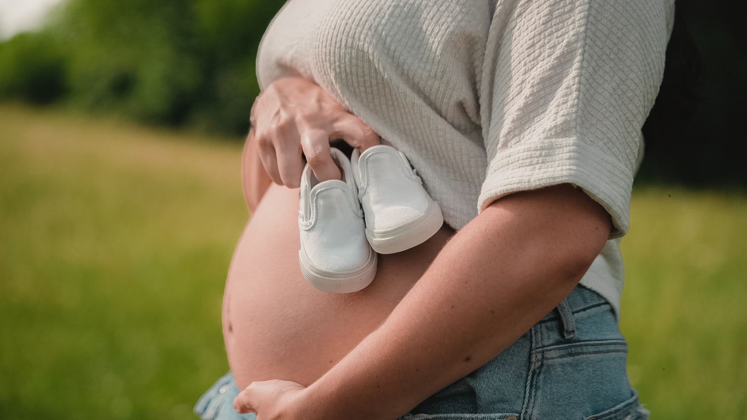 Pregnant woman in jeans holding tiny white baby shoes against her bare belly outdoors in green field.