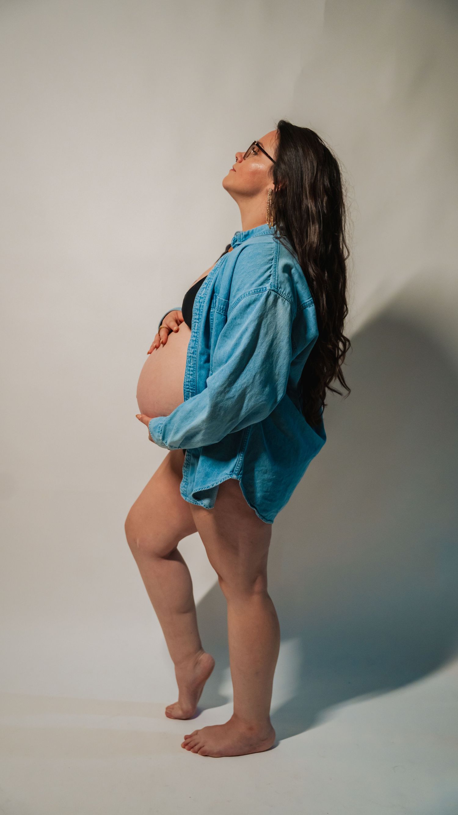 Pregnant woman in open blue denim shirt cradling baby bump, standing barefoot against white background.