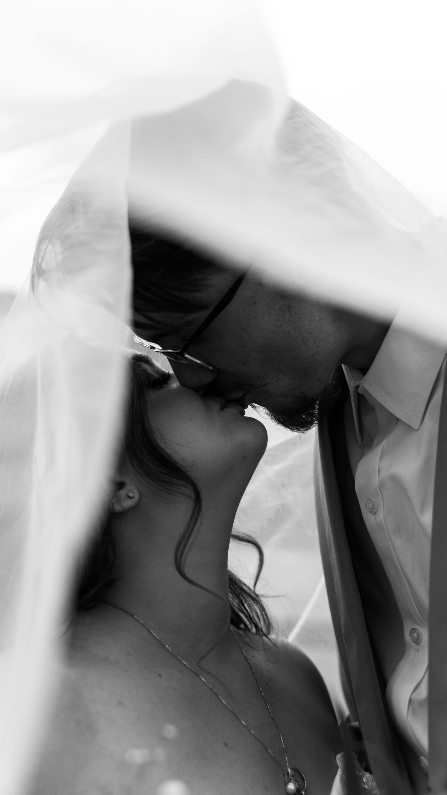 Black and white romantic photo of bride and groom kissing under a flowing wedding veil.