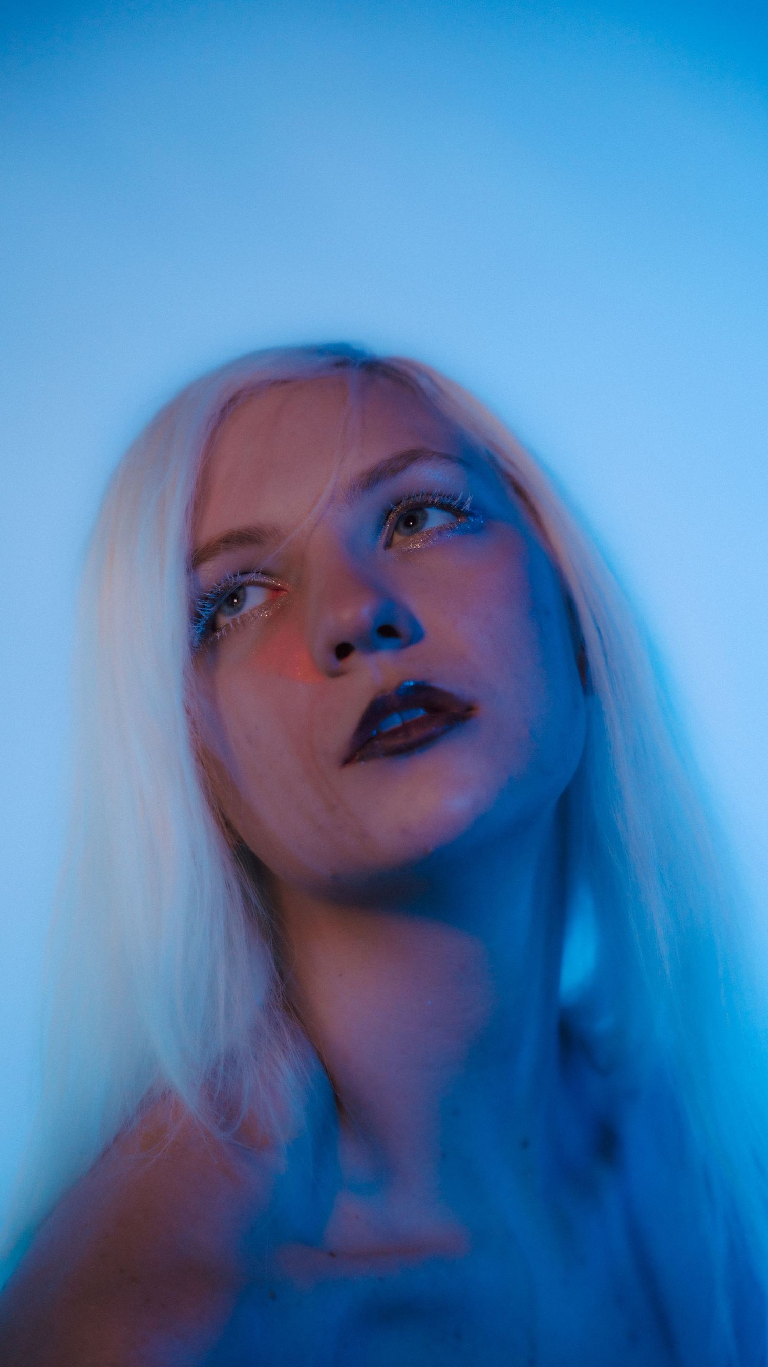 Woman with platinum blonde hair and dark lips poses under dramatic blue and red neon lighting.