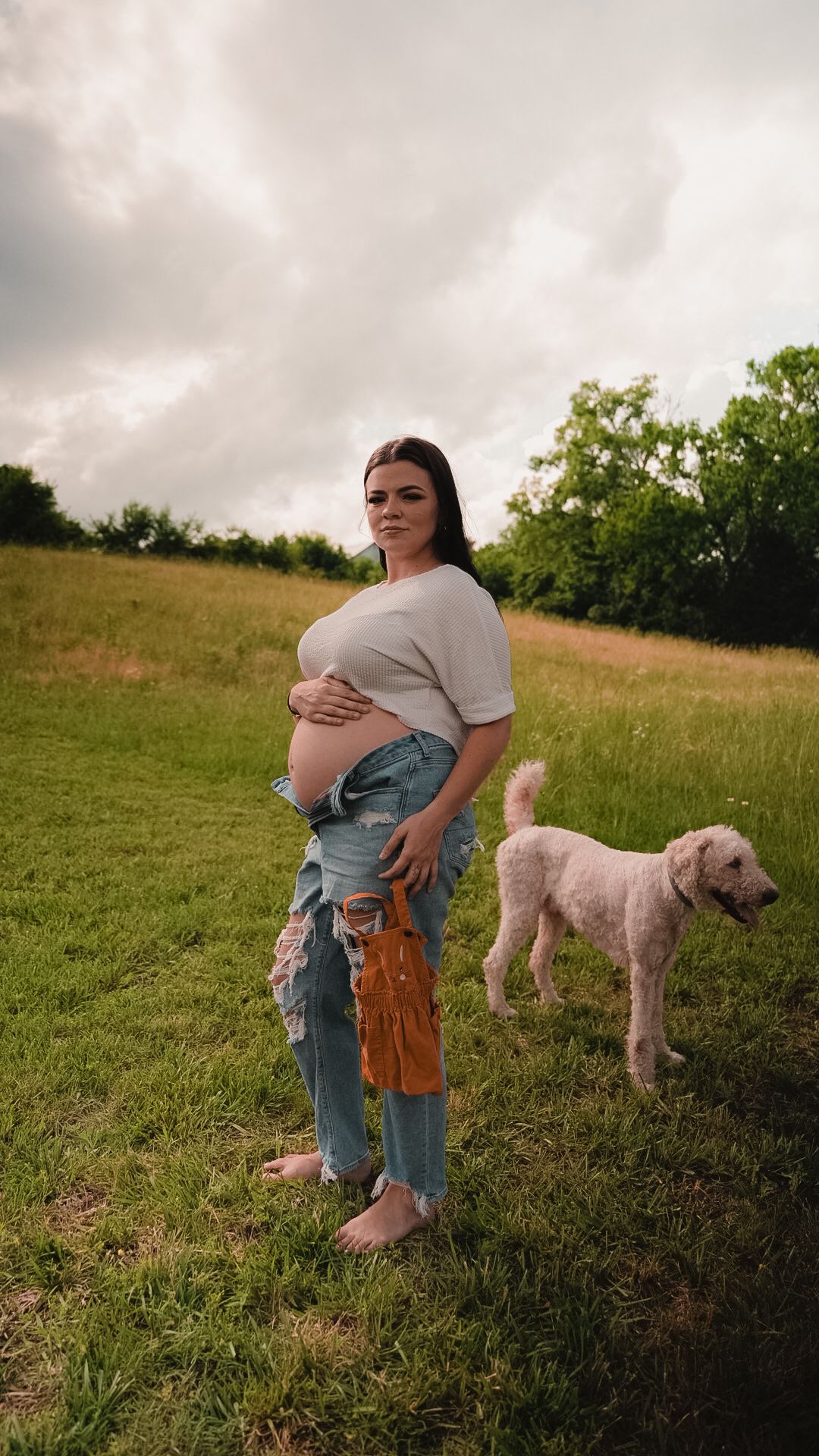 Pregnant woman in ripped jeans holding baby clothes stands beside fluffy dog in grassy field.