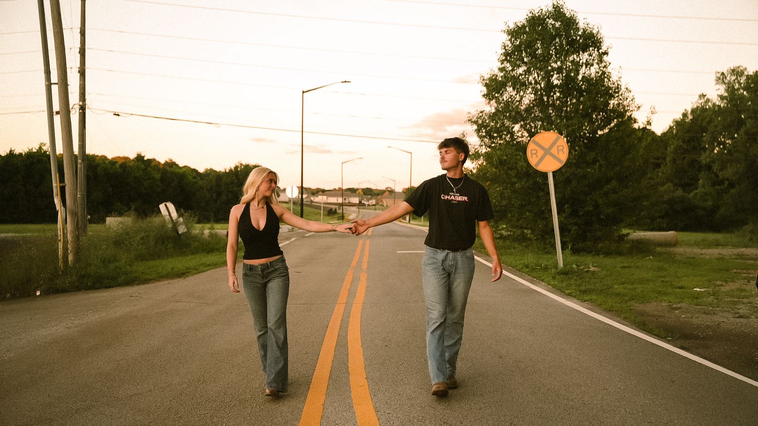 Couple holding hands while walking on a road at sunset, with trees and a railroad crossing sign in the background.