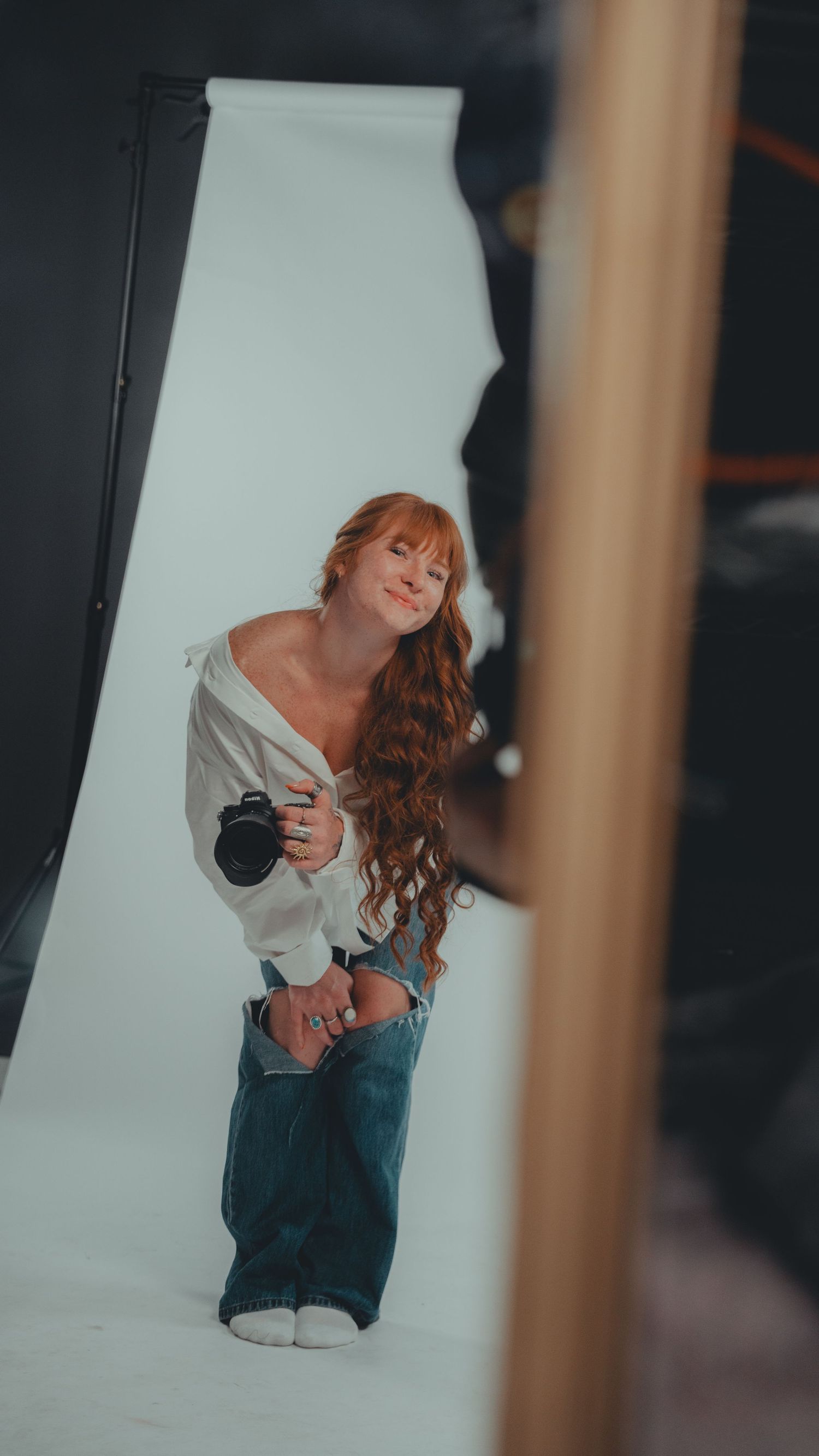 A redhead woman poses in a photography studio holding a camera, wearing an off-shoulder white top and baggy jeans.