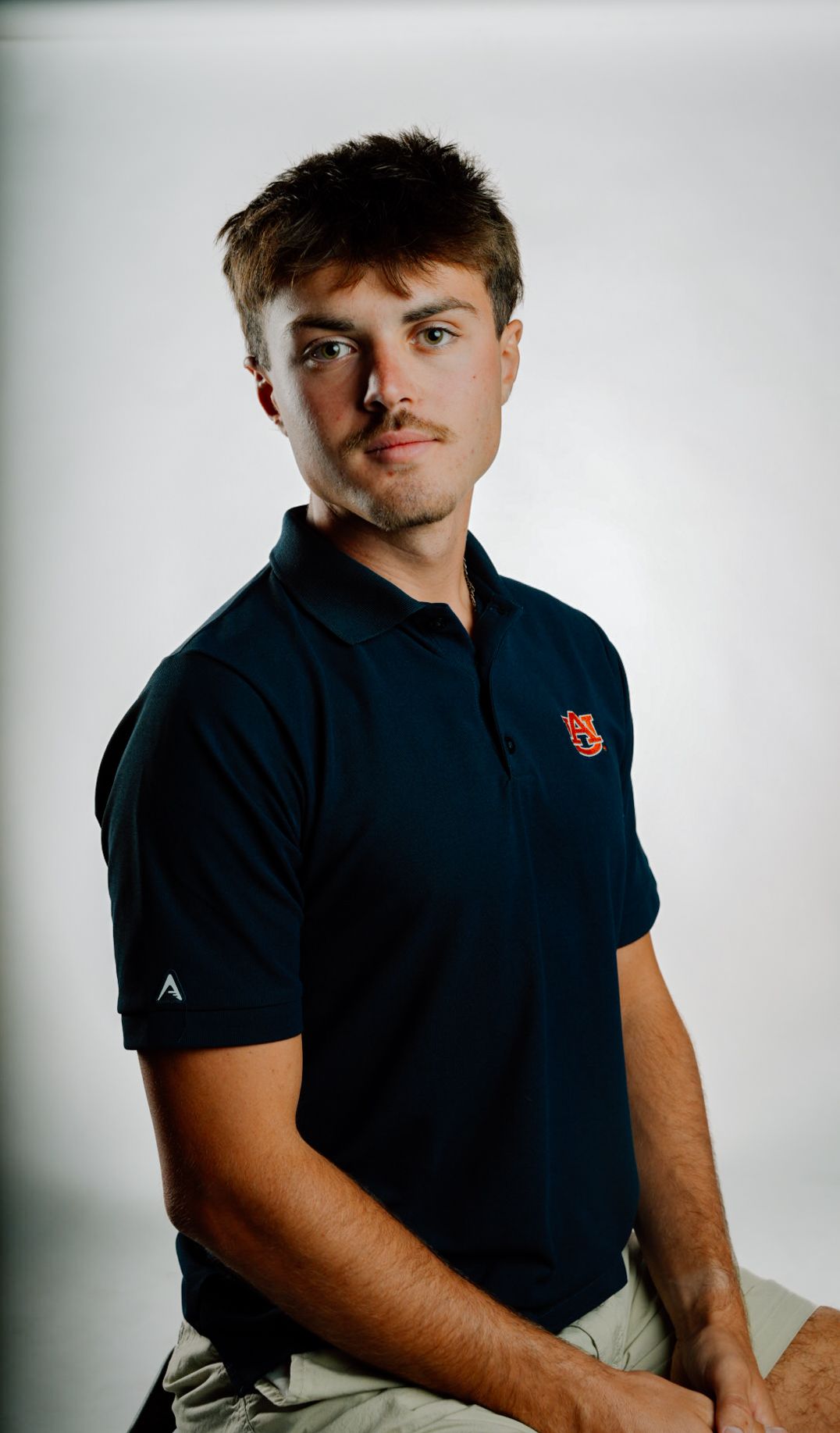Young man in navy blue polo shirt with red logo poses for a professional portrait against white background.