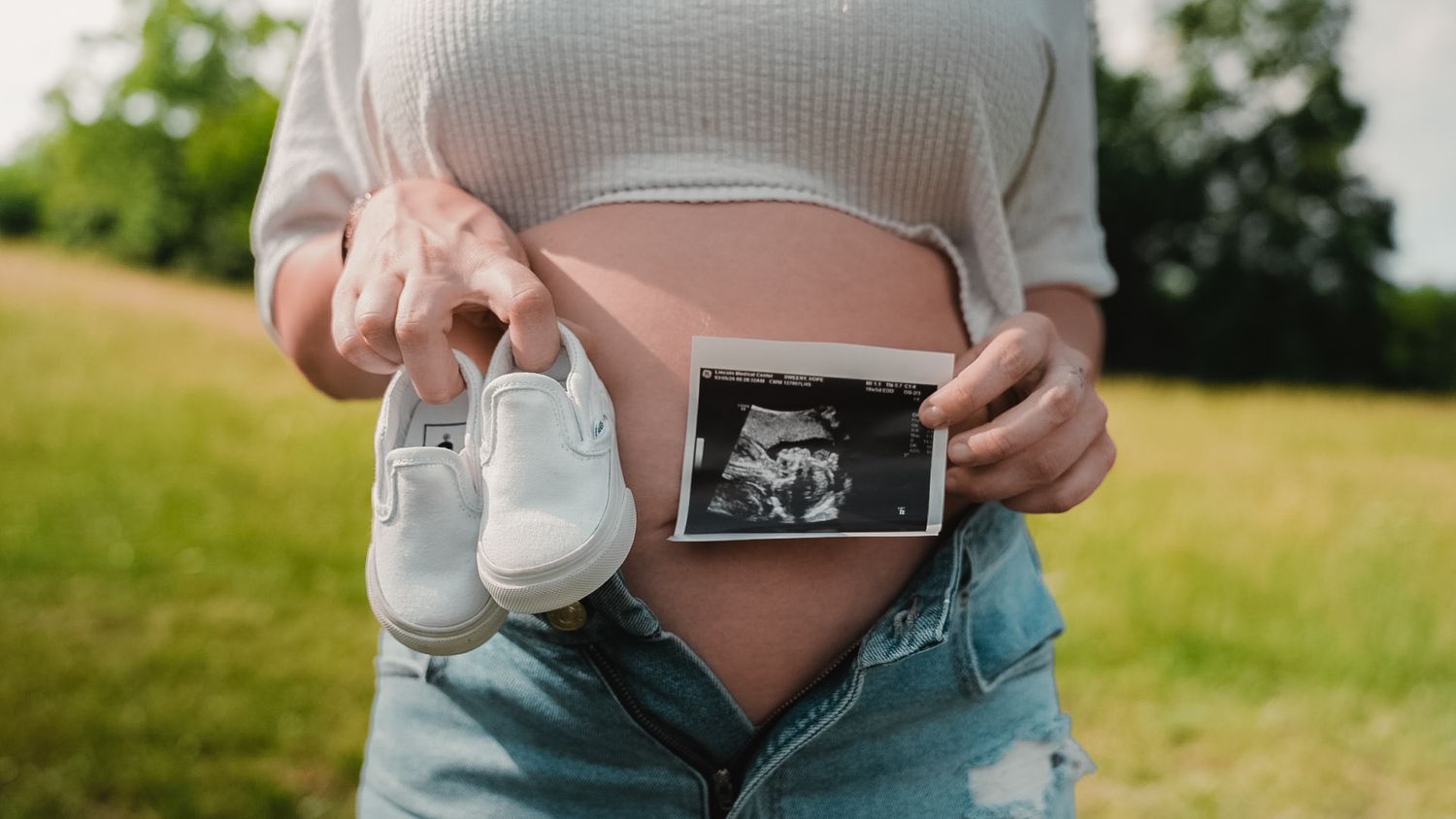 Pregnant woman outdoors holding baby shoes and ultrasound scan photo against her baby bump in a park.