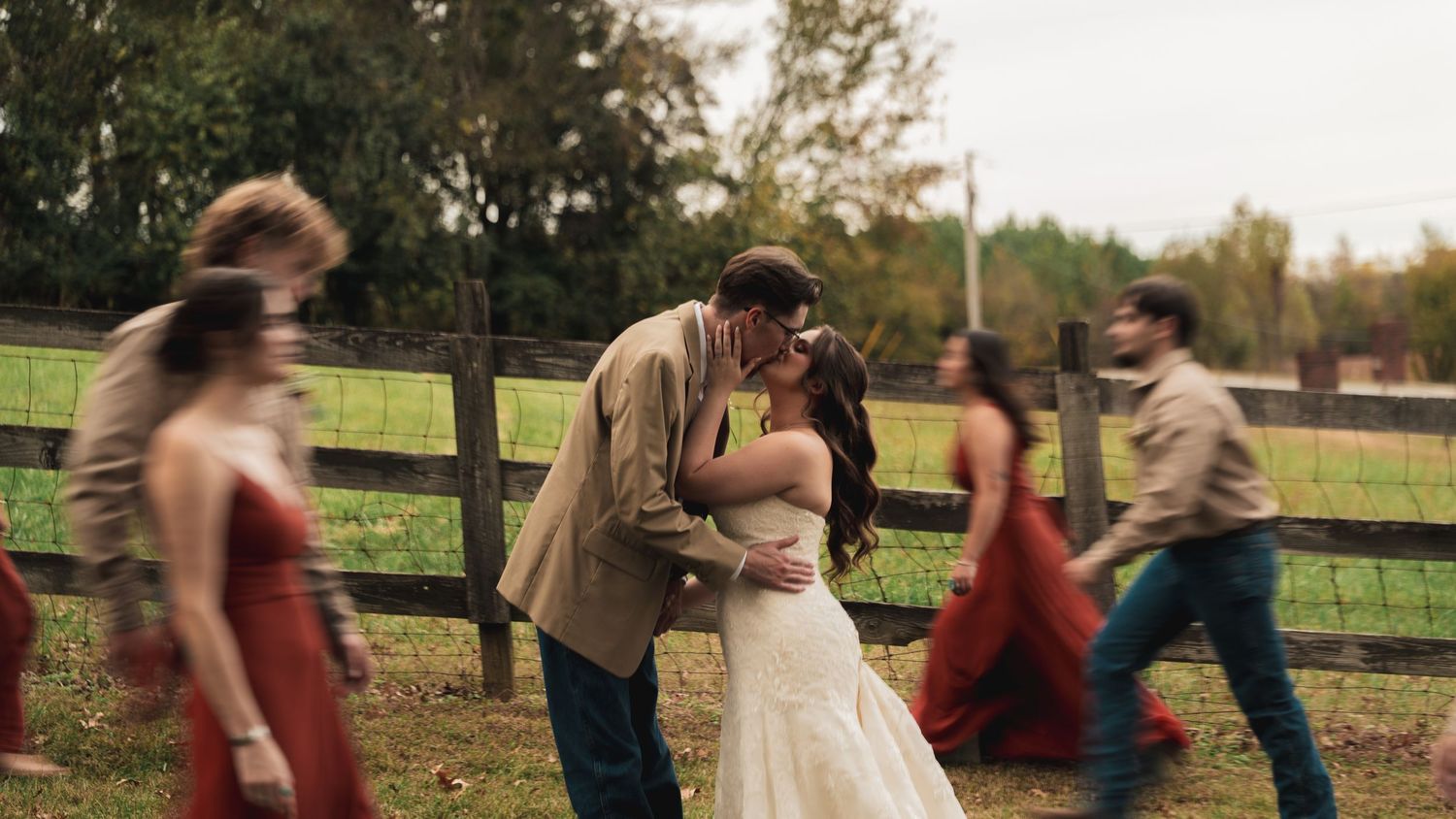 A couple shares a romantic kiss in wedding attire outdoors by a wooden fence, with guests in red dresses walking past.