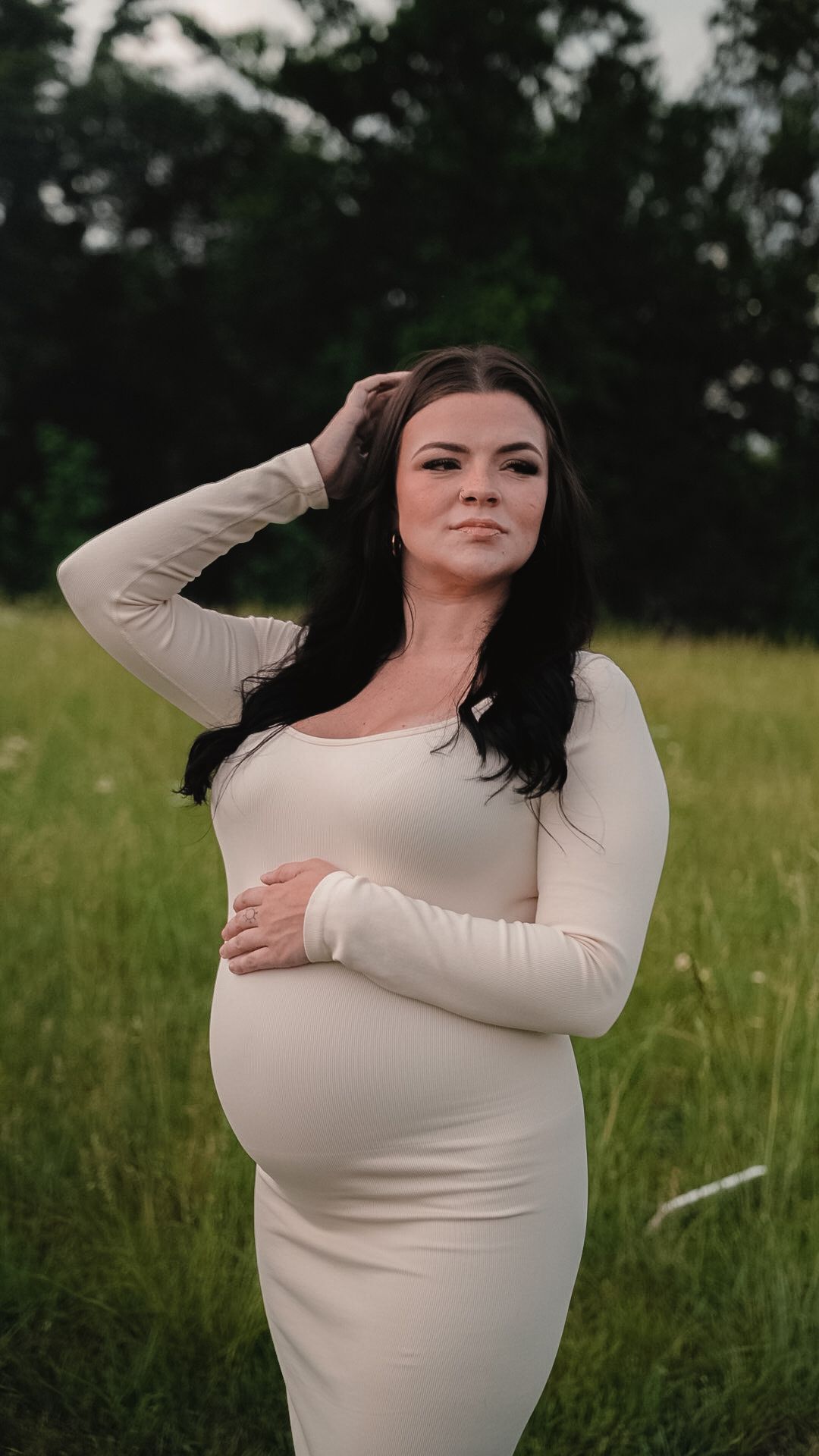 Pregnant woman in fitted white ribbed dress standing in a field, posing with hand in hair.