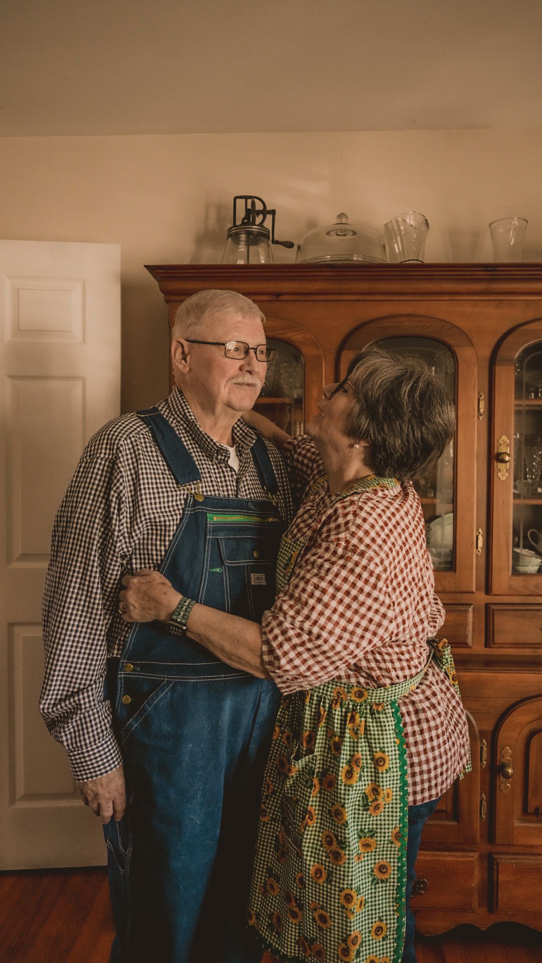 Elderly couple sharing a tender moment indoors beside a wooden china cabinet.