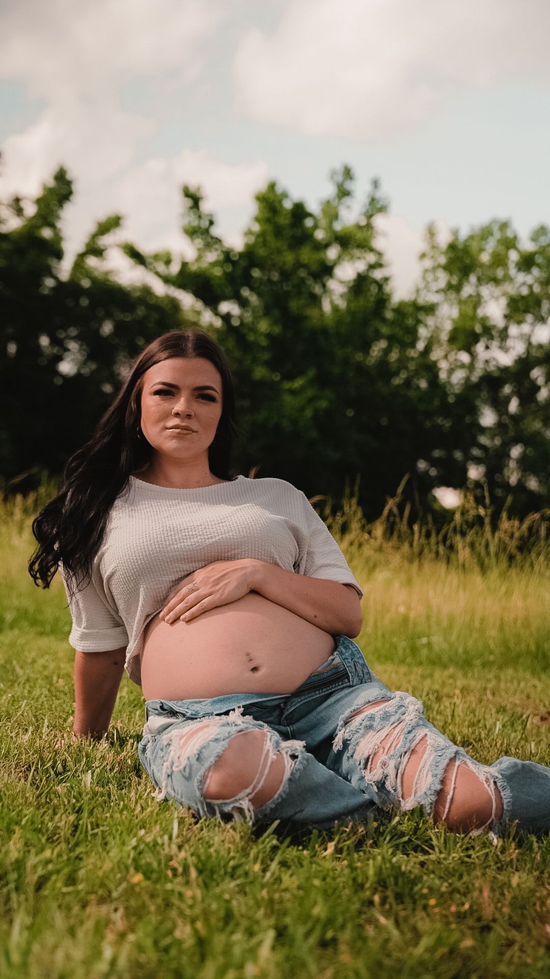 Pregnant woman sitting on grass in ripped jeans and white top, cradling her baby bump outdoors.