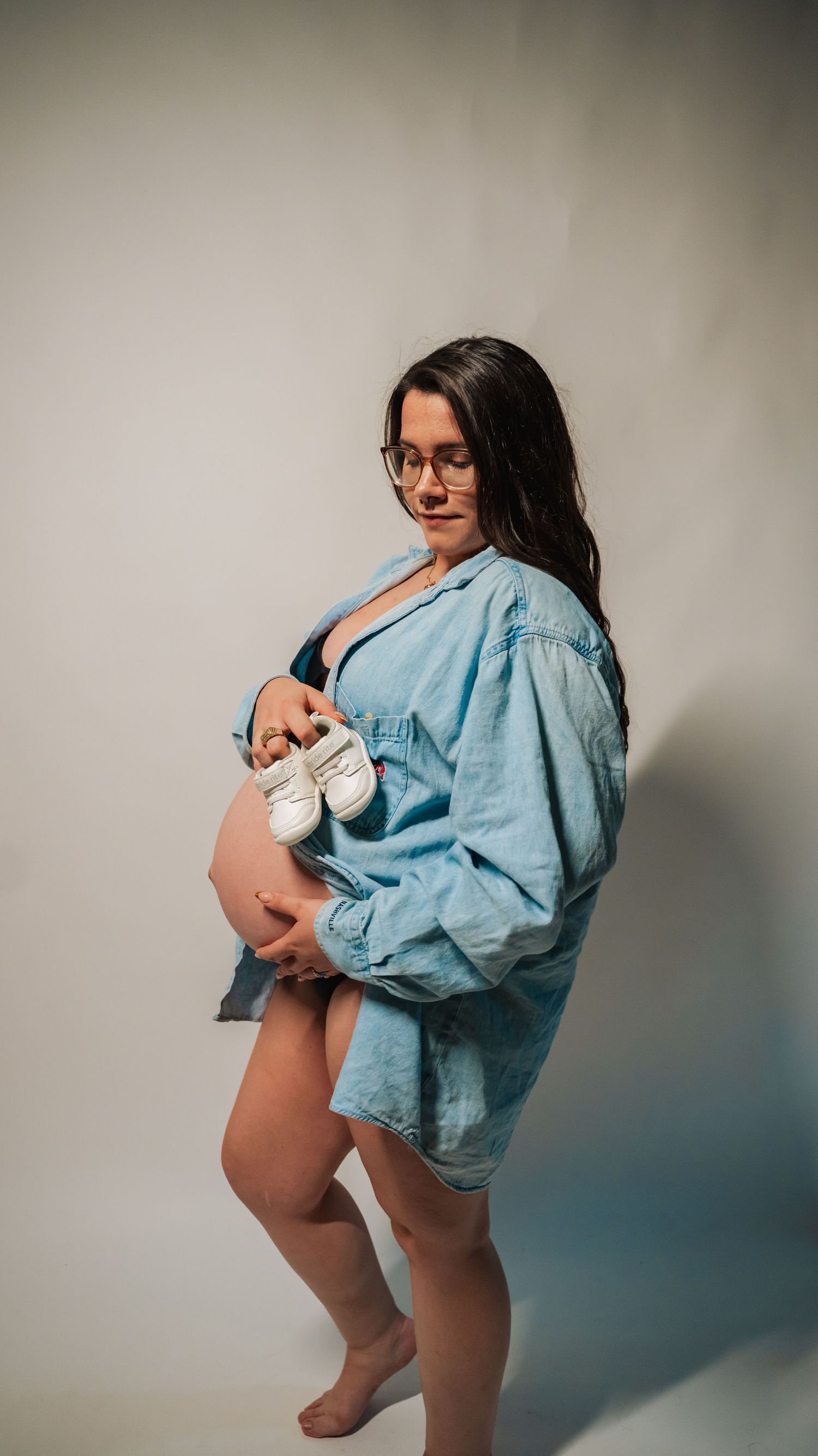 Pregnant woman in denim shirt holding baby shoes and bare bump, maternity photo studio portrait.