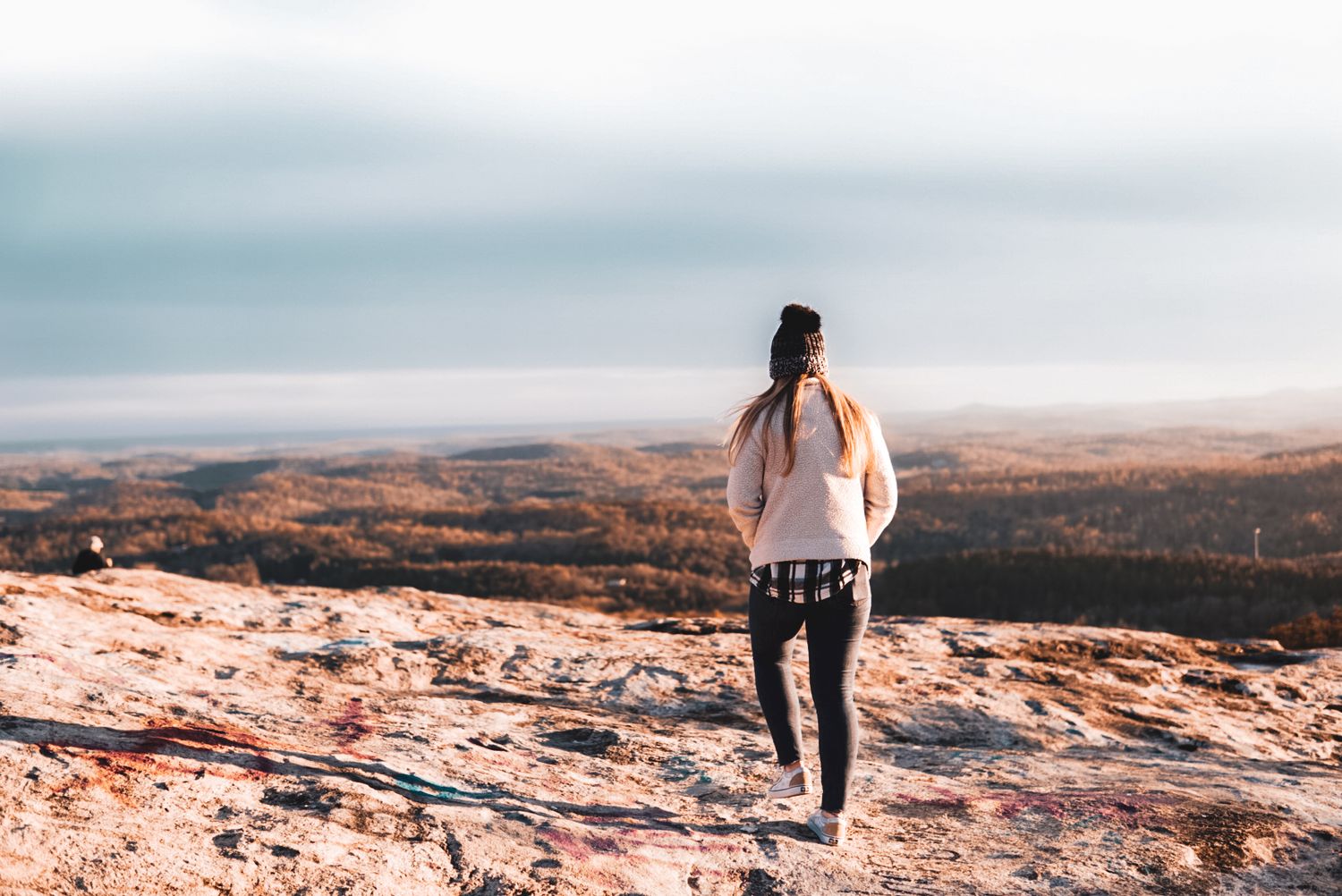 Woman standing alone on a rocky mountain summit overlooking a vast scenic landscape.