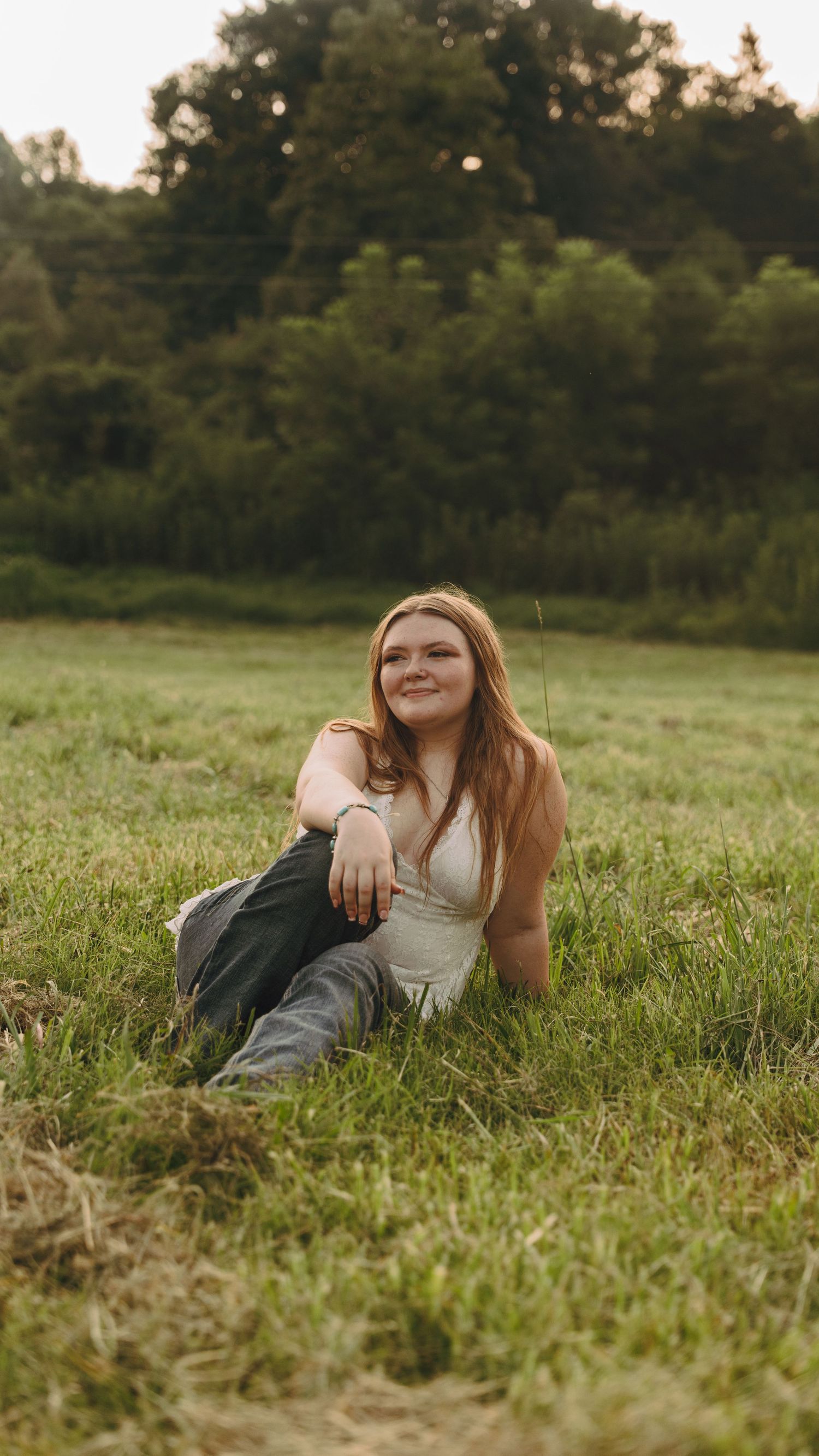 Young woman with long red hair sitting in a grassy field, smiling, wearing a white top and jeans.