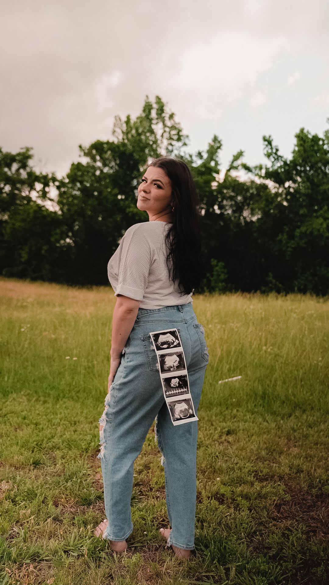 Woman in jeans displaying ultrasound photos on back pocket, standing in green field for pregnancy announcement.