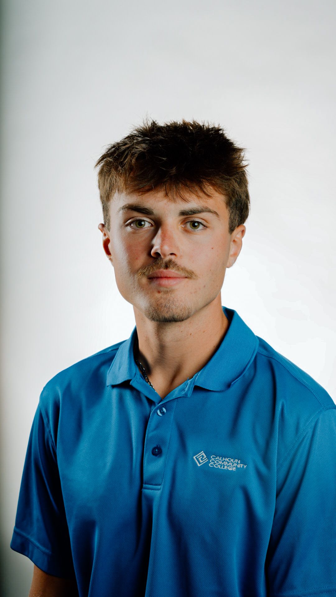 Young man with light brown hair wearing blue Calhoun Community College polo shirt posing against white background.