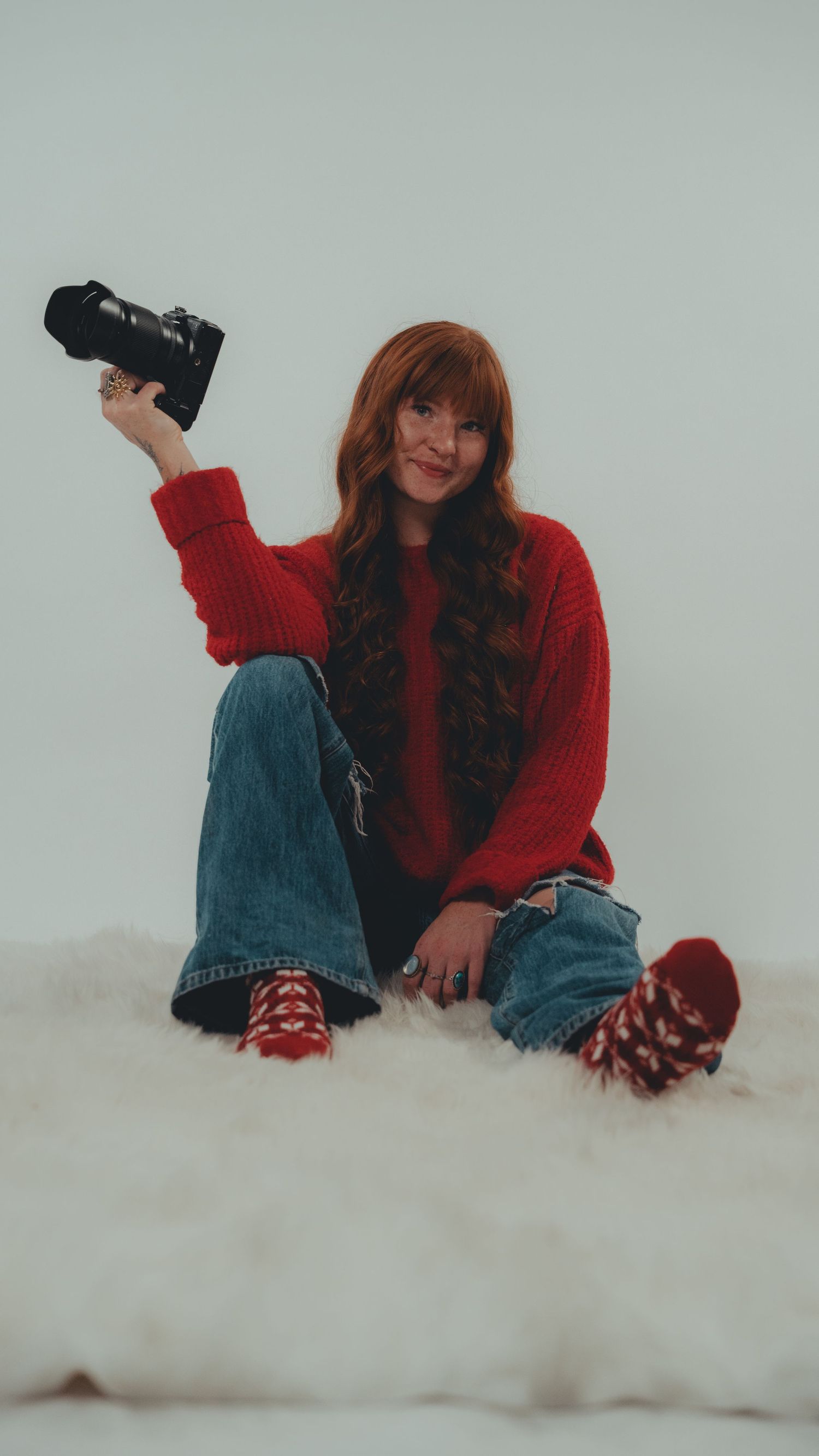 A smiling redhead woman in a red sweater and jeans sits on a fluffy rug, holding a camera up in the air.