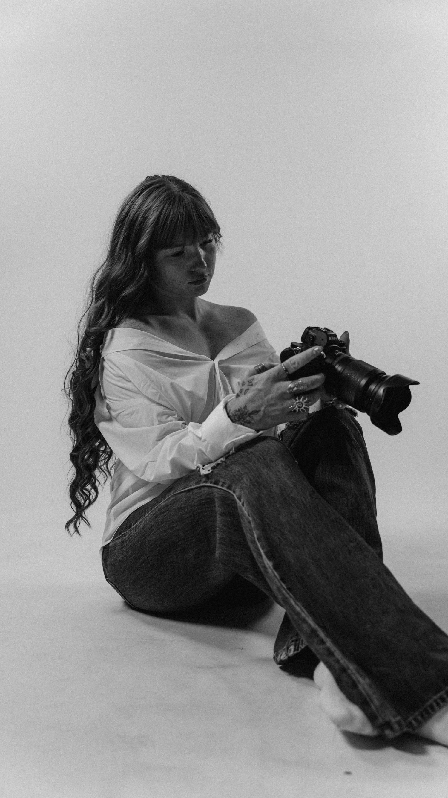 Black and white photo of woman sitting on floor holding DSLR camera in white shirt and jeans.