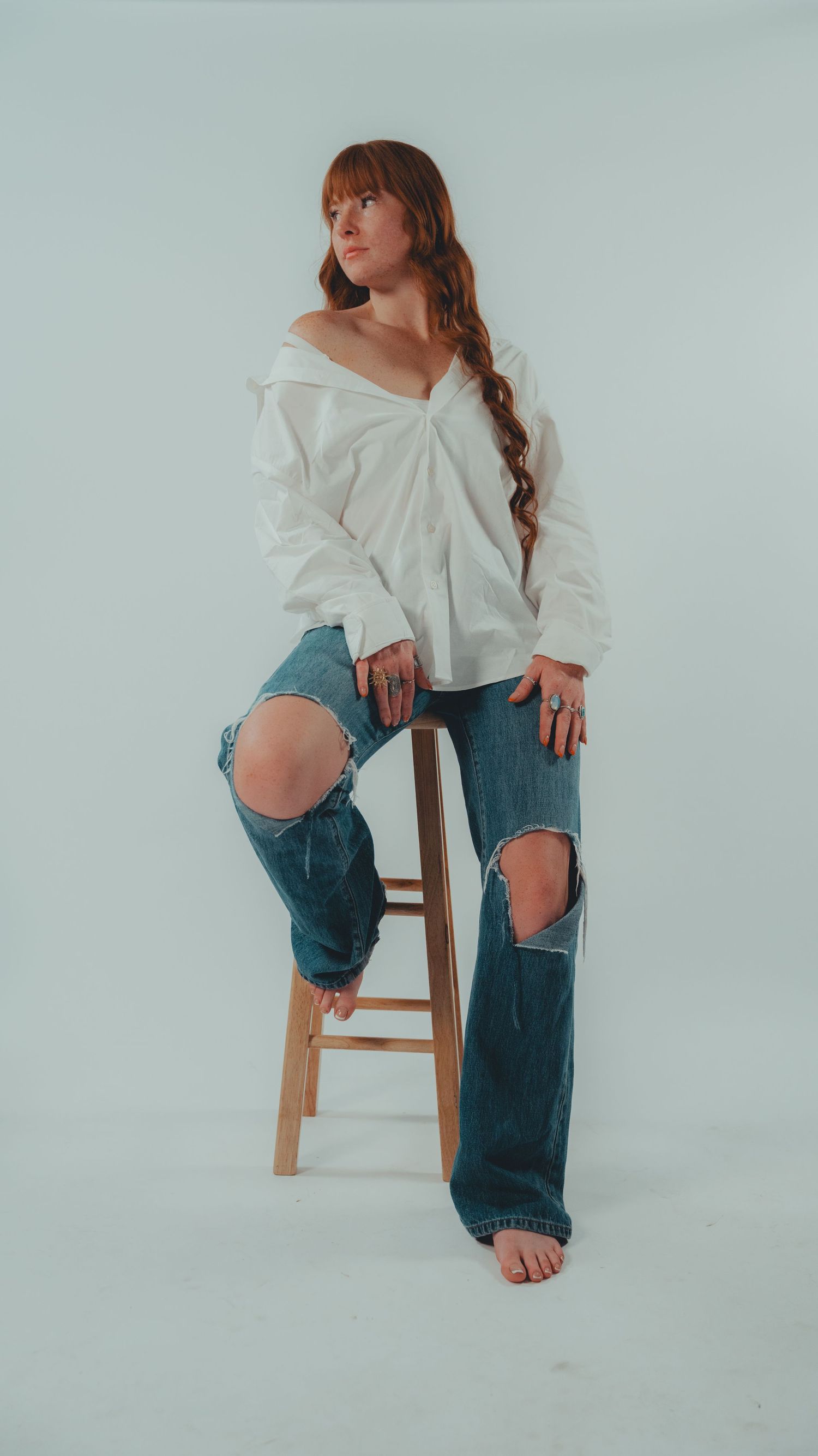 Woman in white off-shoulder top and ripped blue jeans poses on wooden stool against light gray background.