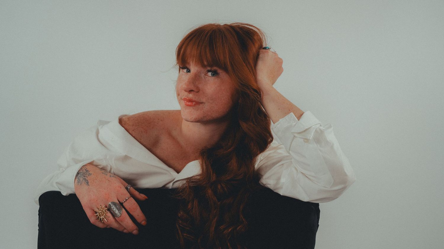 Young woman with auburn hair poses elegantly in white off-shoulder blouse against light gray background.