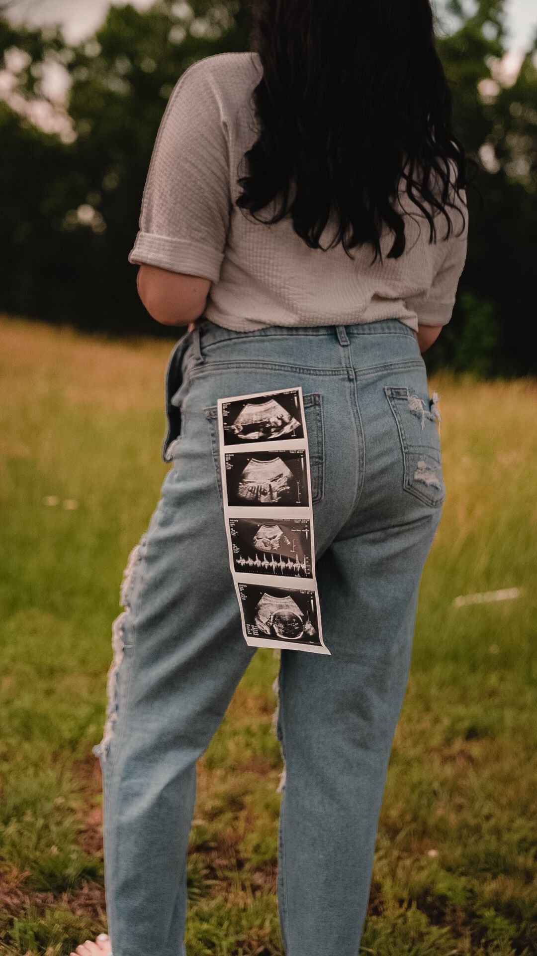 Pregnant woman in light blue jeans standing in field, displaying ultrasound photos tucked into back pocket.