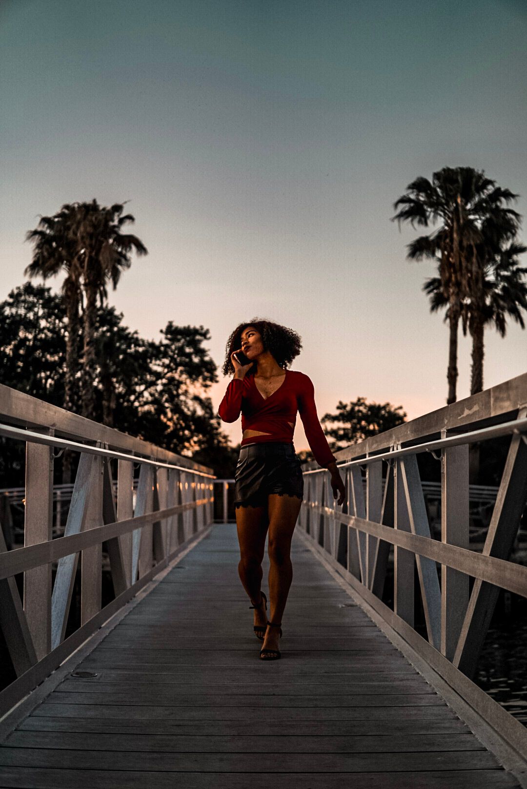 Woman in red top and black skirt walking on wooden bridge at dusk with palm trees.