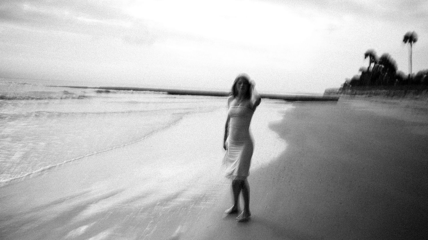 Black and white photo of woman in white dress standing on beach near ocean waves.