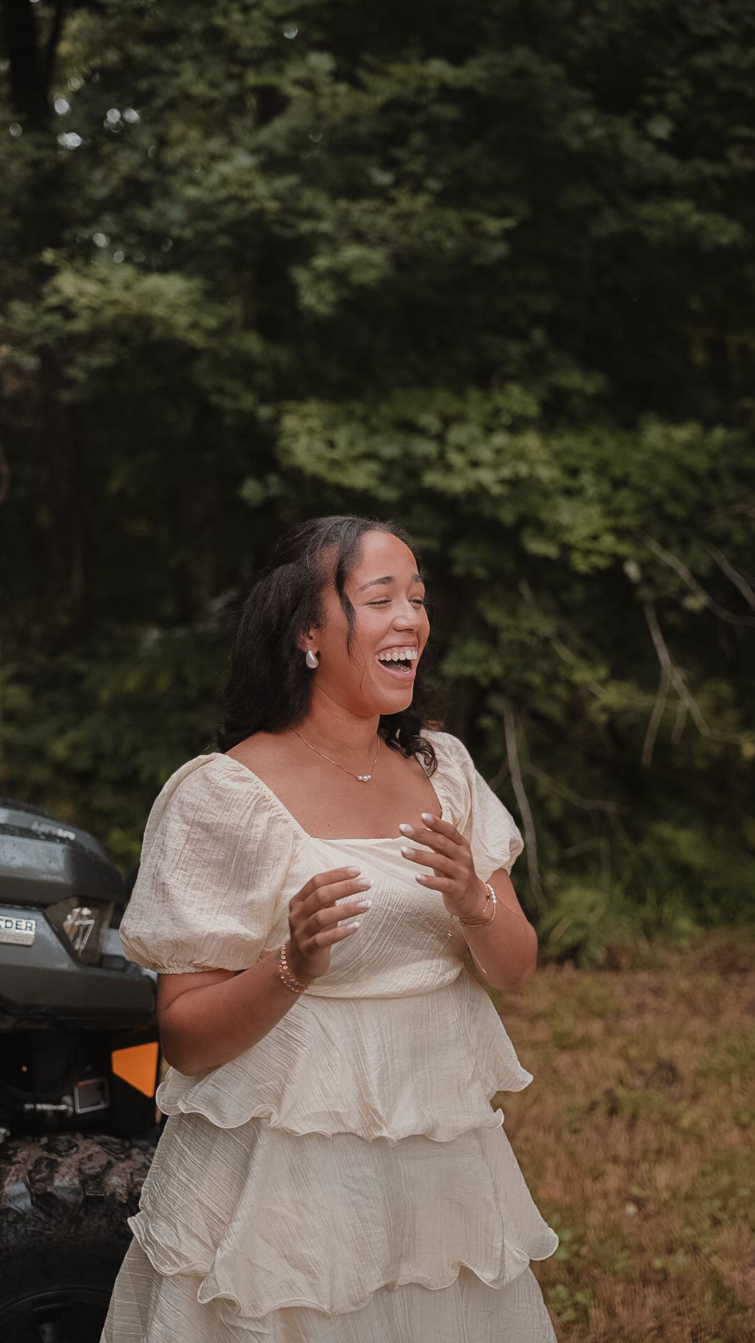 A joyful woman in a white dress laughs outdoors near an ATV surrounded by lush green trees.