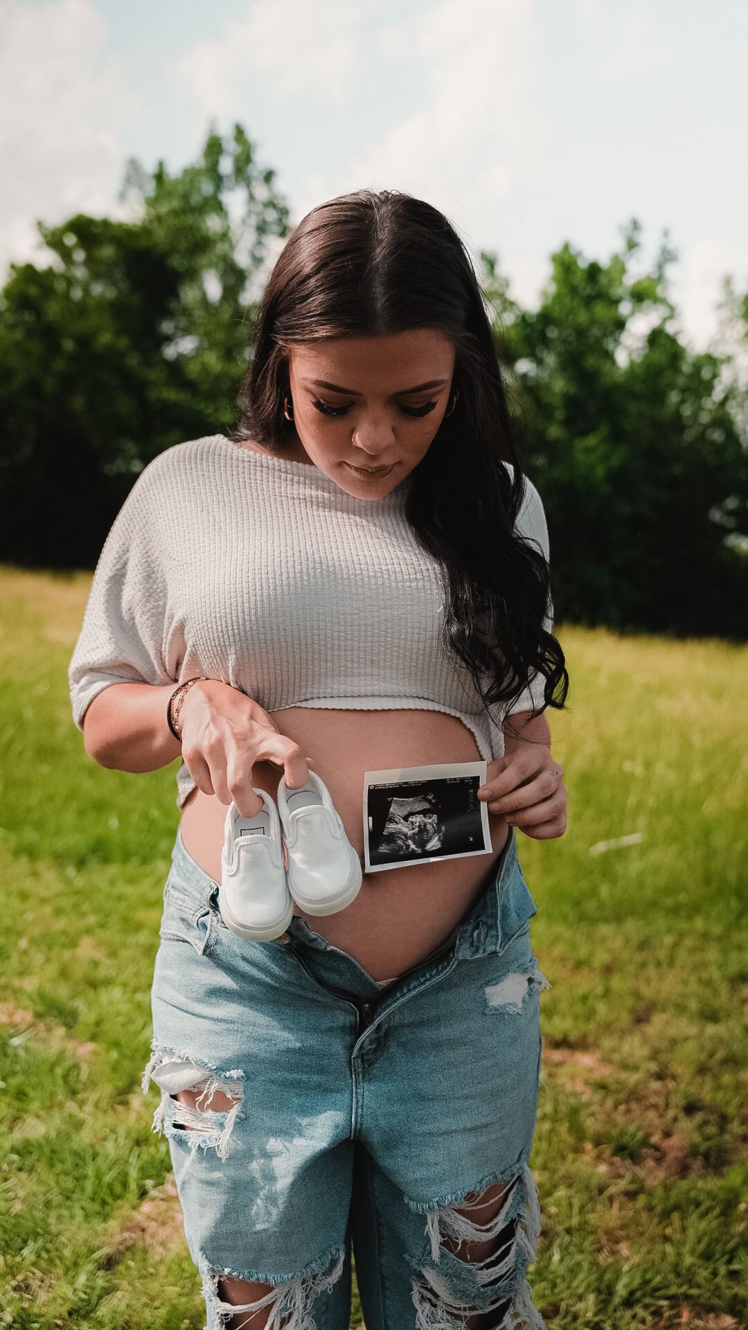Expectant mother in ripped jeans displaying ultrasound image and tiny white shoes on her pregnant belly outdoors.