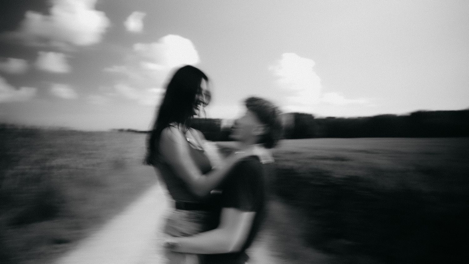 Black and white photo of a couple embracing in a field, motion blur adding romantic energy to the outdoor scene.