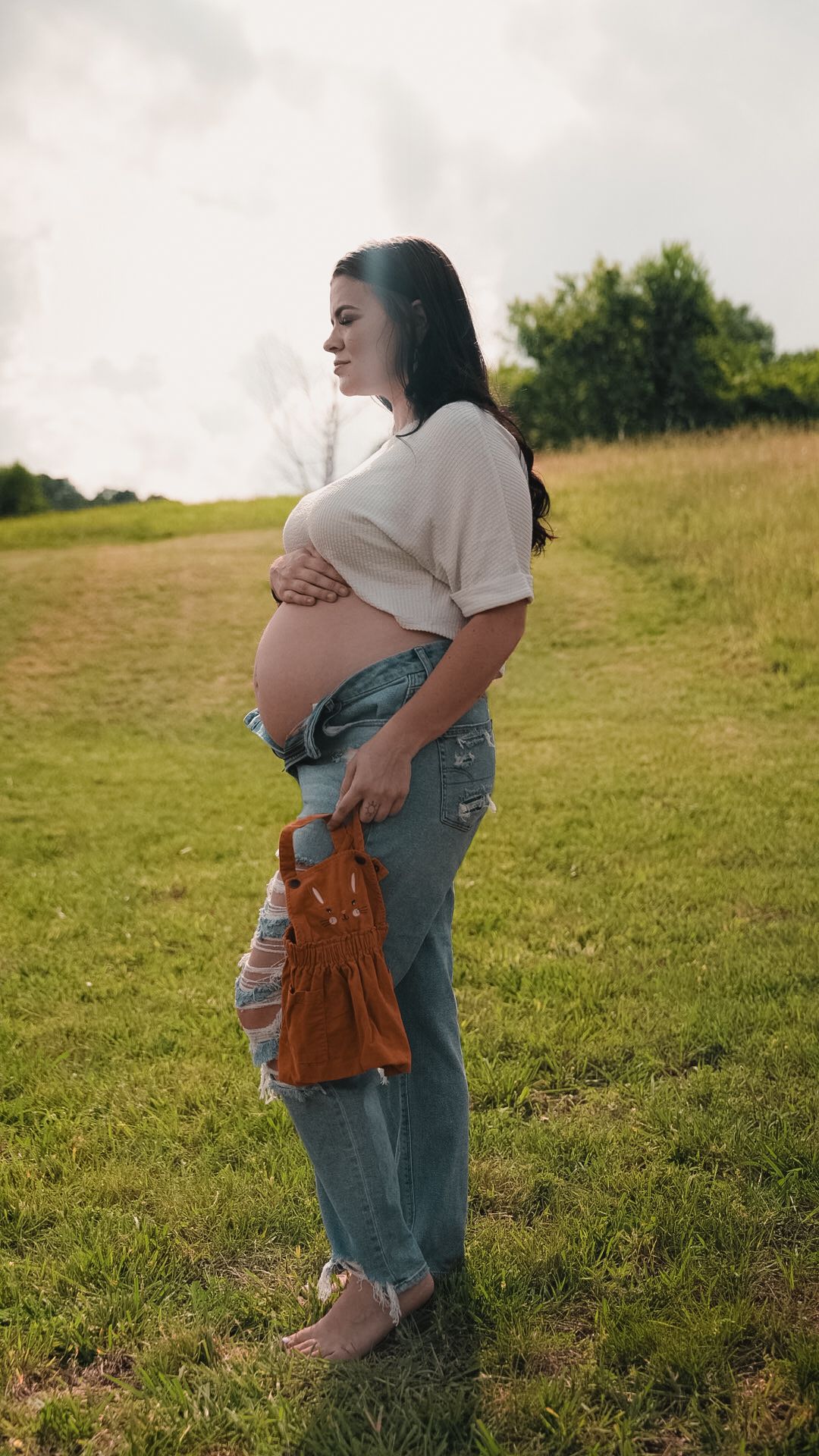 Pregnant woman in white crop top and ripped jeans cradling baby bump outdoors in green field.