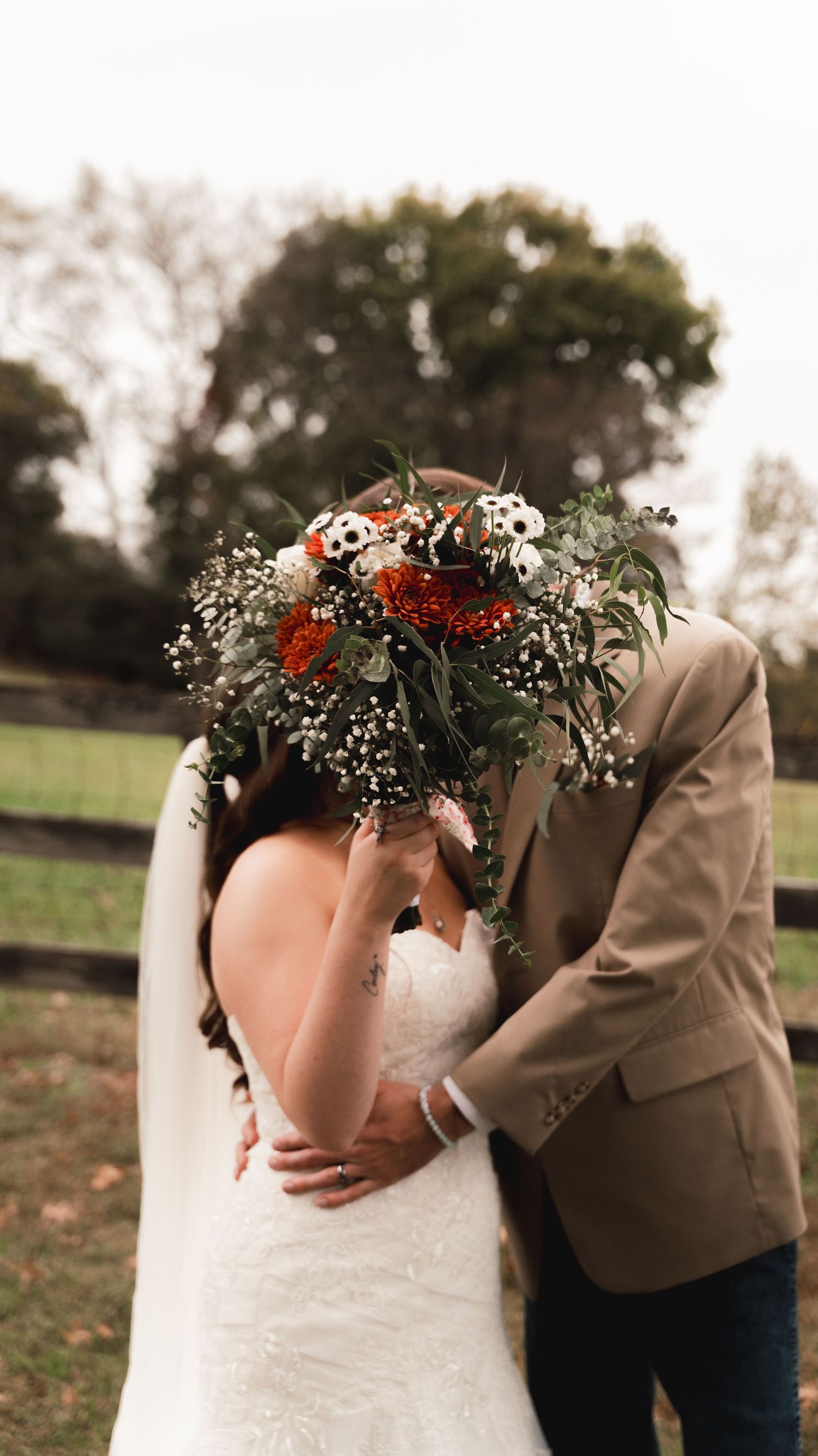 A bride and groom kiss behind a lush bridal bouquet of red, white flowers and greenery at an outdoor fall wedding.