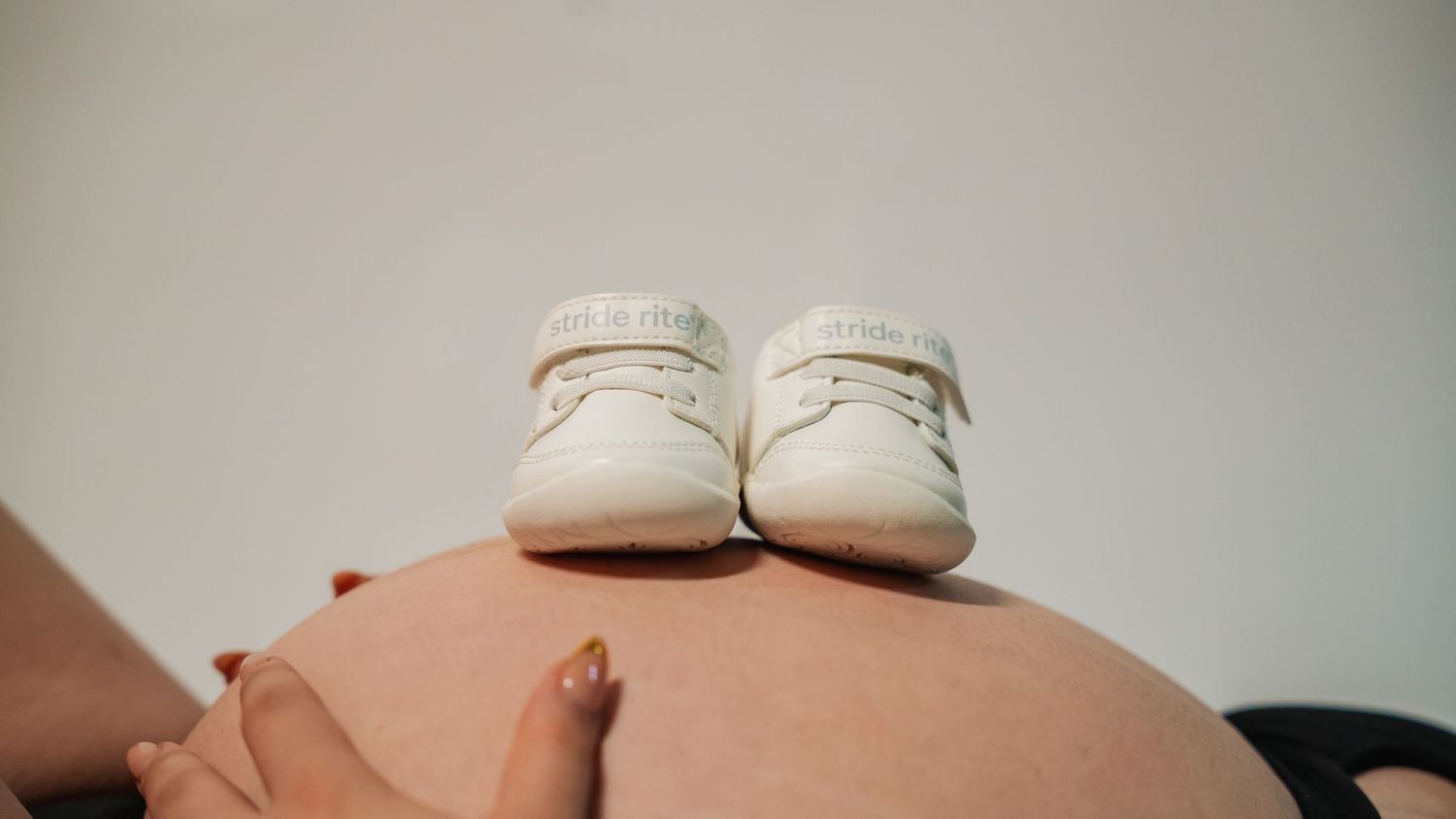Pregnant woman's bare baby bump with tiny white baby shoes resting on top against a light background.