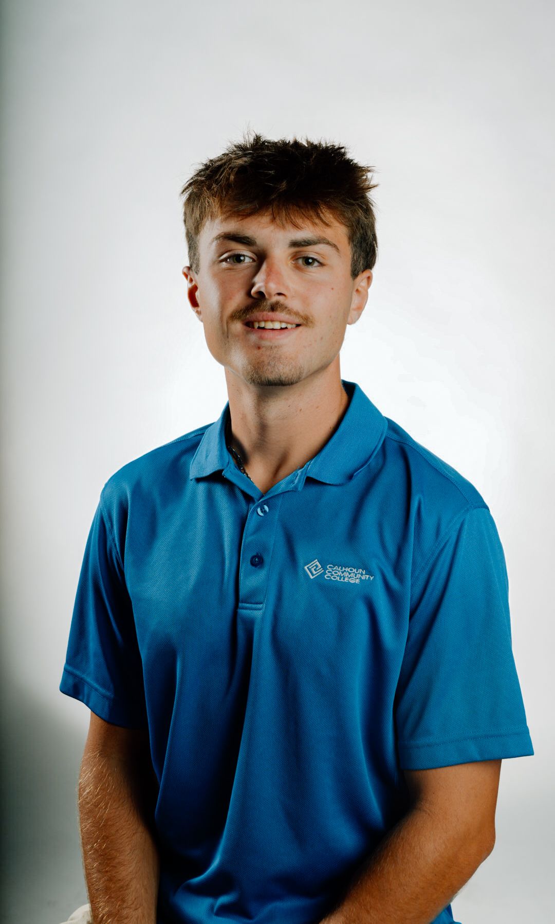 Young man smiling in a blue polo shirt with a college logo on a white background.