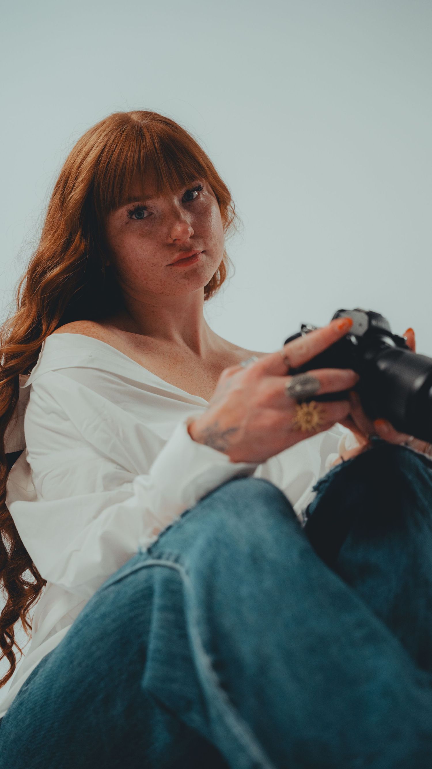 Redhead woman with freckles holding a DSLR camera, wearing white off-shoulder top and blue jeans.