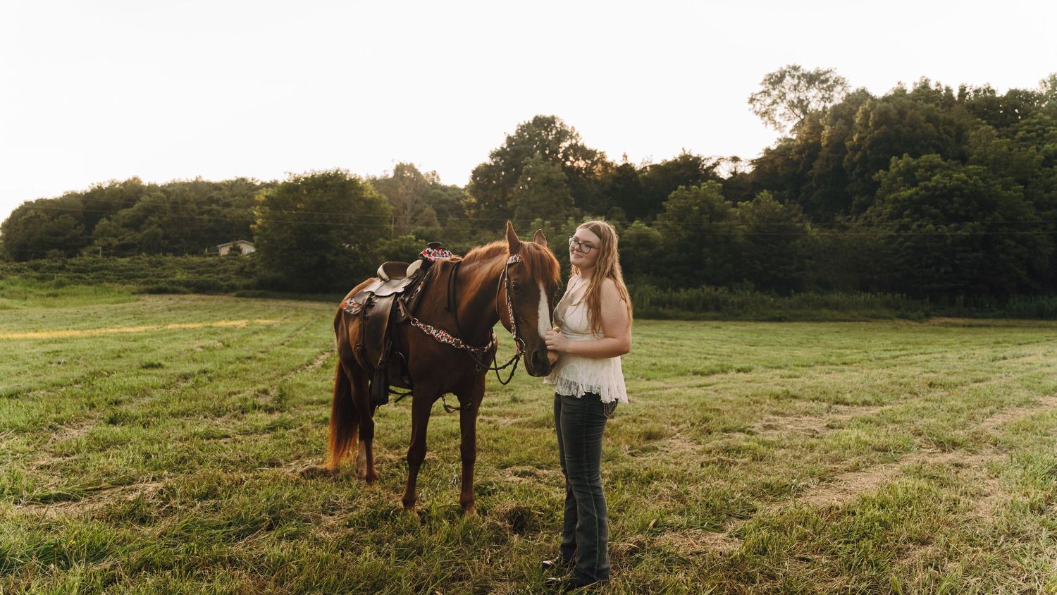 A young woman stands smiling beside a saddled brown horse in a lush green pasture with trees in the background.