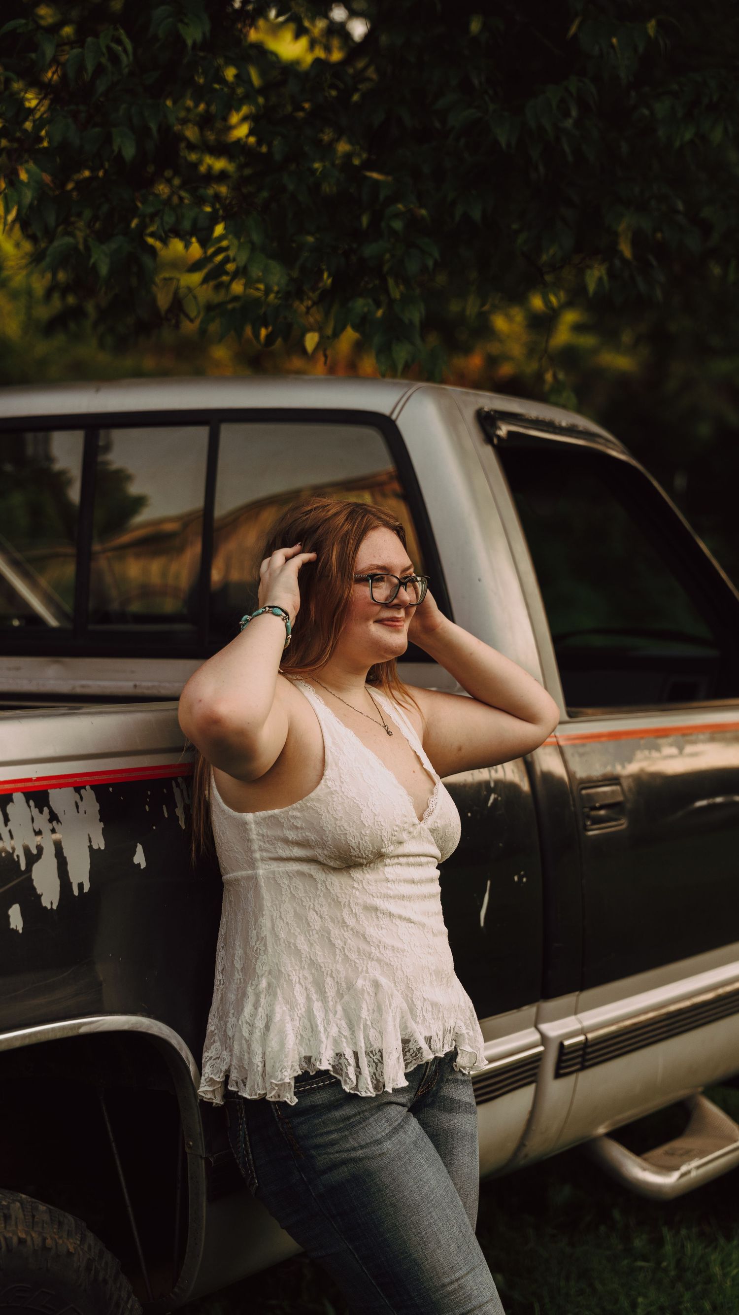 Young woman with glasses in white lace top and jeans posing against a vintage pickup truck outdoors.