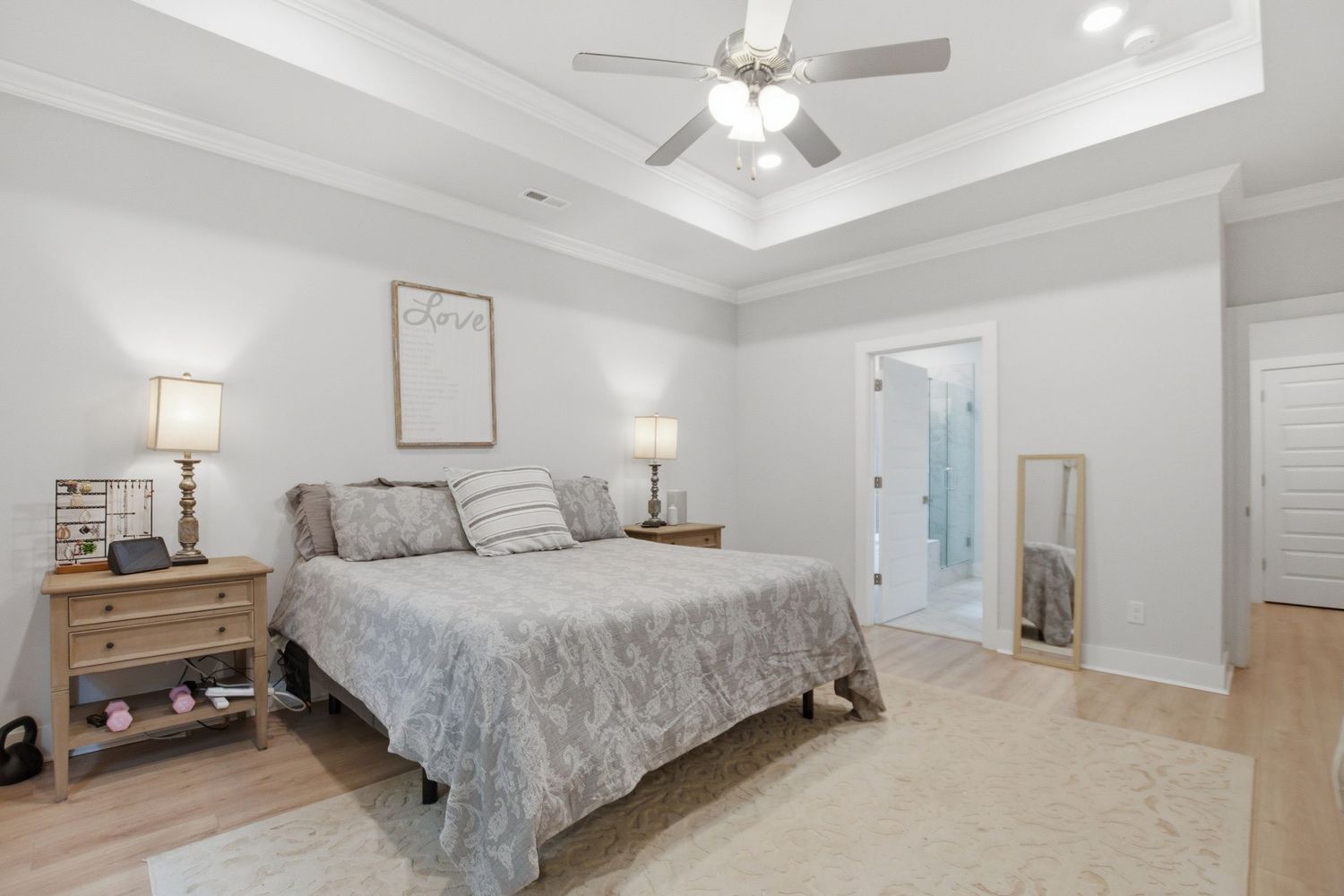 Bright master bedroom with tray ceiling, ceiling fan, hardwood floors, and en-suite bathroom in a modern home.