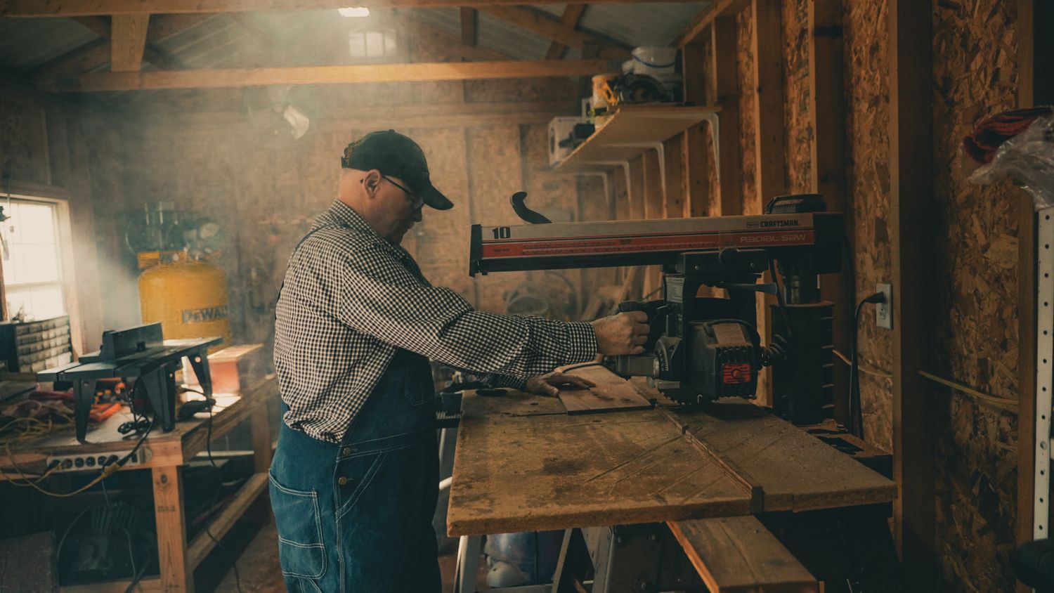 Man working with wood at a workbench inside a rustic workshop with shelving.